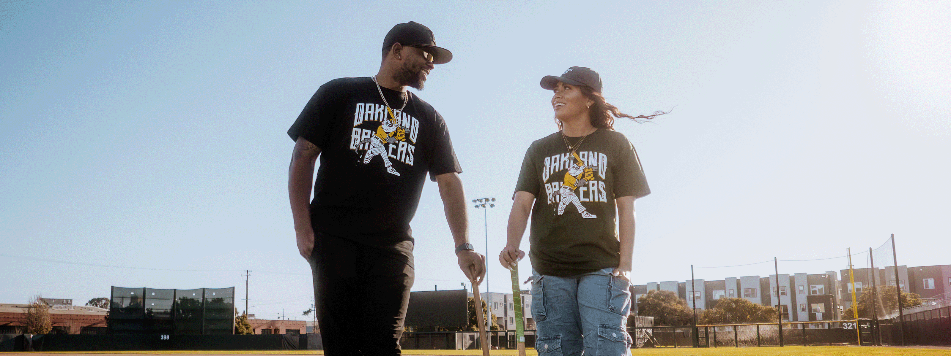 Two people in matching "Bakken Bearcats" shirts and caps walk on a baseball field under a clear sky, holding bats. They're smiling at each other, with stadium lights and buildings visible in the background.