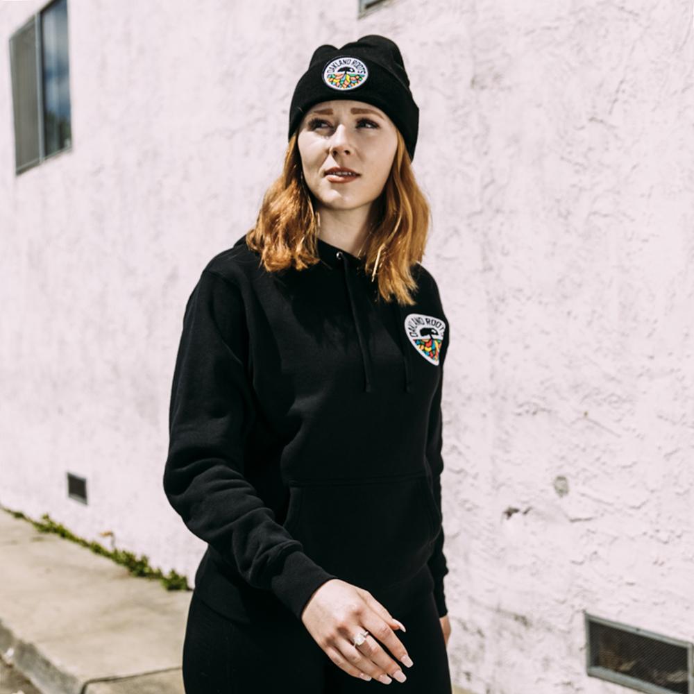 A person with shoulder-length, light brown hair stands outdoors against a white wall. They wear a black hoodie and an Oakland Roots SC Cuff Beanie, both featuring a colorful circular logo. The sunlight casts shadows on the wall. The person looks slightly to the left, with one hand by their side.