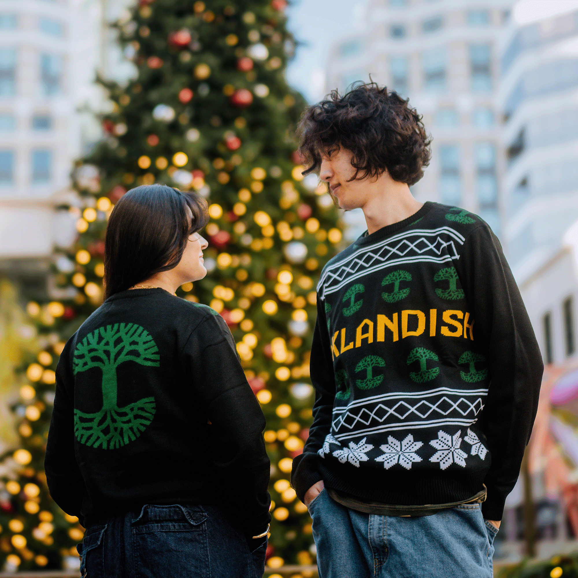 Two people stand outside facing each other, wearing black knit sweaters with green patterns. One sweater prominently displays the word "Oaklandish" with snowflakes; the other showcases a green tree design. A large, decorated Christmas tree and tall buildings blur in the background, highlighting their festive Oaklandish 2024 Ugly Sweaters.