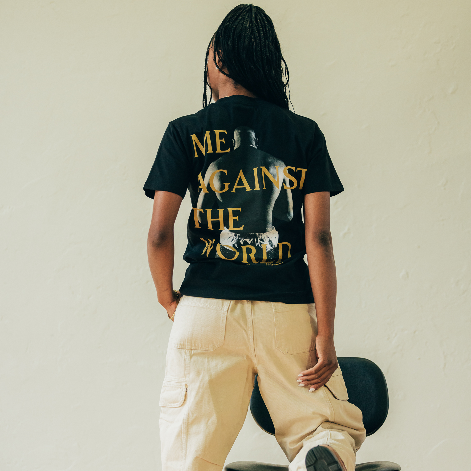 A person with braided hair, channeling Tupac's iconic style, faces away while wearing the "Against the World Tee" by Oaklandish. The black T-shirt features gold text that declares "ME AGAINST THE WORLD." They sit on a black chair against a simple backdrop, dressed in beige cargo pants. The soft and natural lighting enhances the hip-hop vibe of the scene.