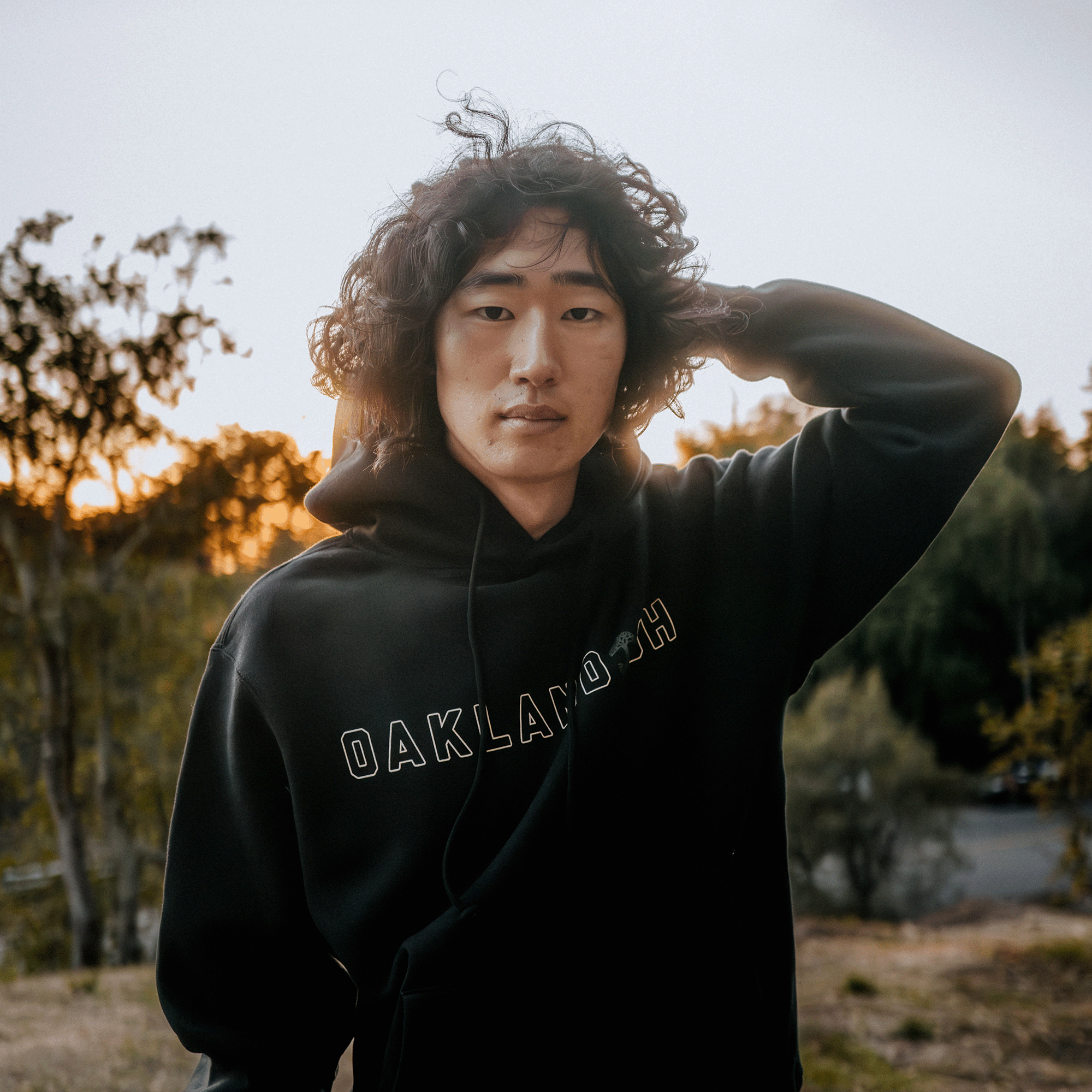 A person with curly hair wearing a black Bandit Hoodie by Oaklandish stands outdoors as the sun sets. The hoodie, designed for men, features the "OAKLANDISH" logo. They gaze directly at the camera with one hand lightly touching their head, set against a backdrop of blurred trees and a gently glowing sky.