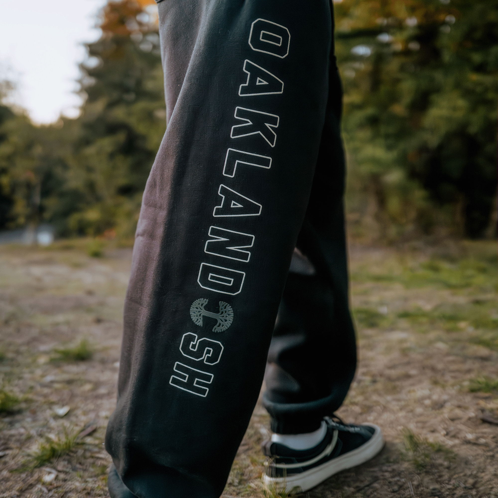 A close-up of a person wearing Bandit Sweatpants from Oaklandish, showcasing "OAKLAND" in bold white letters with "HGS" underneath, designed for men's sizing. The sweatpants feature a hidden zipper pocket for added practicality. In the background, a blurred forest path complements the individual's black and white sneakers.