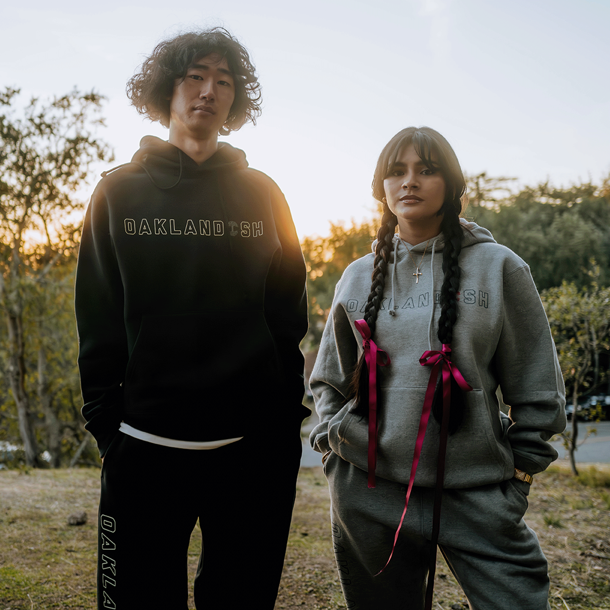 As the sun sets outdoors, a man and woman enjoy the serene atmosphere, each wrapped in cozy "Oaklandish" attire. The man wears a stylish black Bandit Hoodie with matching sweatpants, while the woman sports a gray set accented with pink ribbons. Trees frame the tranquil scene.