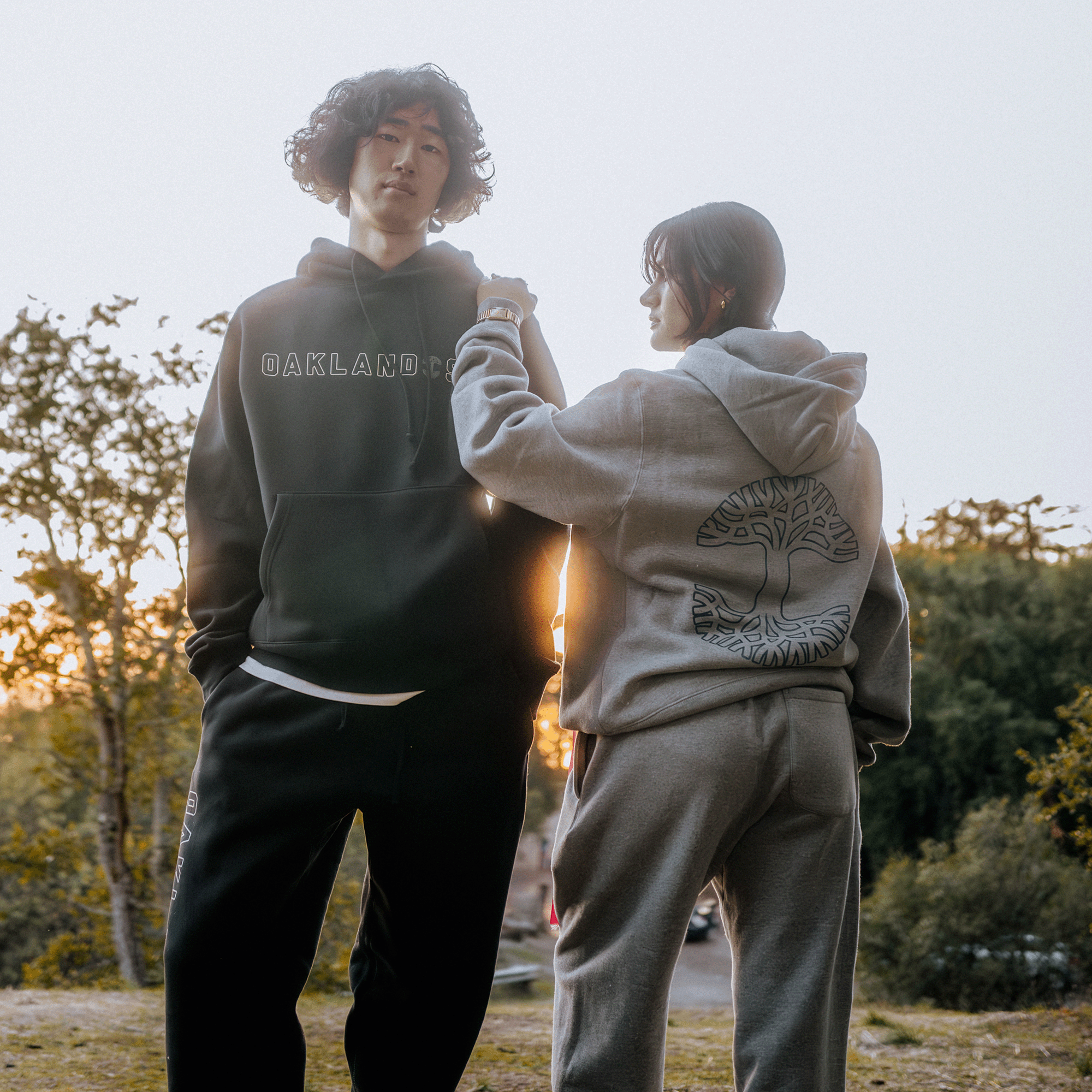 Two individuals clad in hoodies stand outside as the sun sets. On the left, a person dons a dark Oaklandish Bandit Hoodie emblazoned with "Oakland" on the front. Next to them, another person is dressed in a men's heather grey hoodie that showcases a tree design on the back. The scene is enriched by trees and soft sunlight in the background.