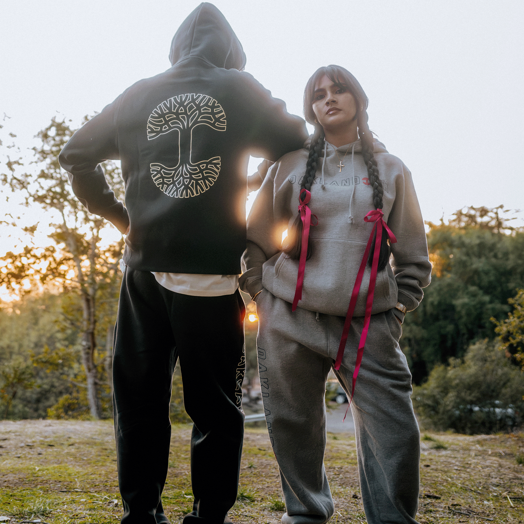 Two individuals outdoors are styled in comfy Bandit Sweatpants and hoodies from Oaklandish. The left person sports a black hoodie featuring a white tree design, while the right showcases a heather grey hoodie with red ribboned braids. Set against a backdrop of trees and sunset, this relaxed vibe fits men's sizing perfectly.