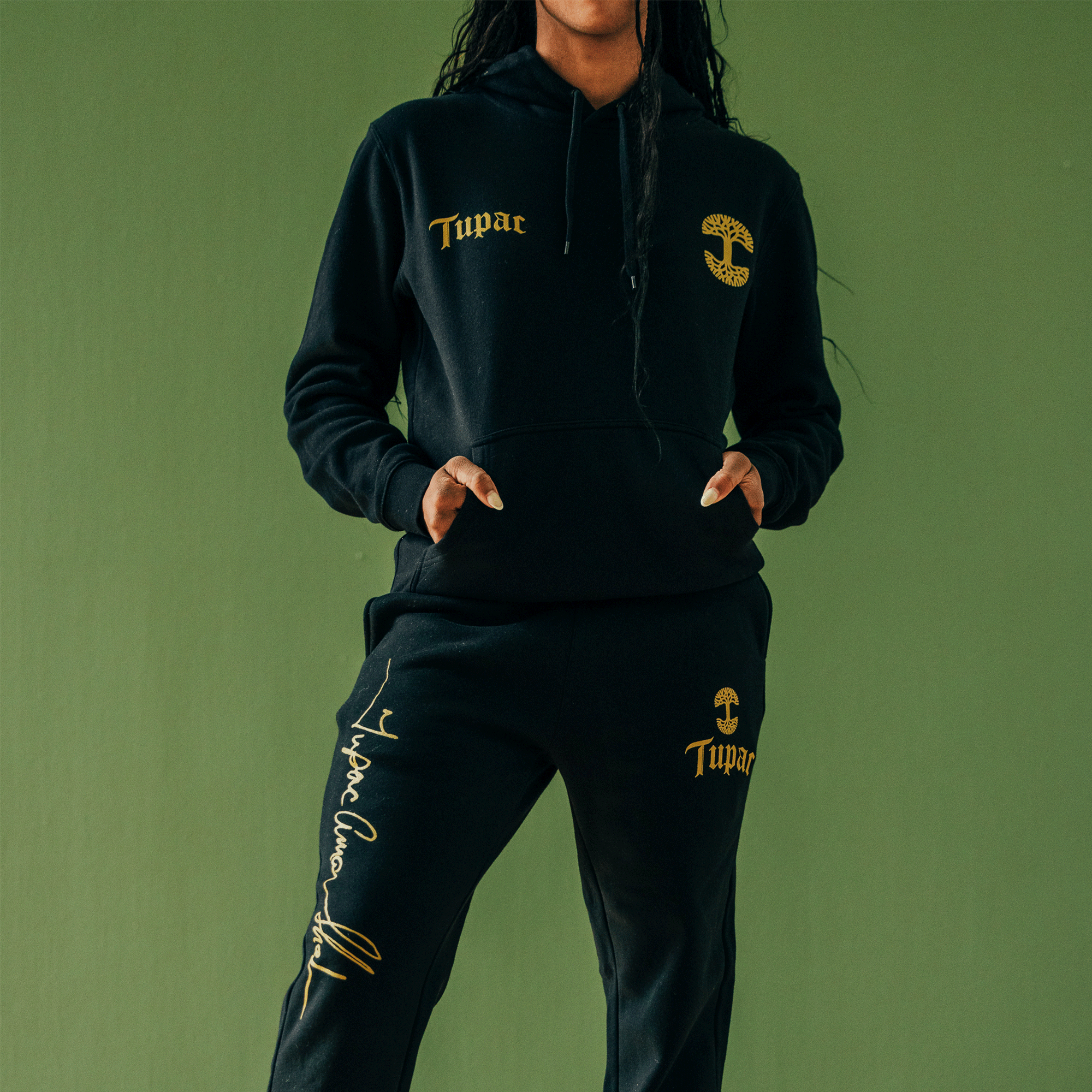 Dressed in a black Bless Jogger set, featuring gold embroidery of the "Tupac" name and stylized cross, this Oaklandish x Tupac piece exemplifies cultural influence against a green background. The joggers prominently display "Tupac" along with his signature.