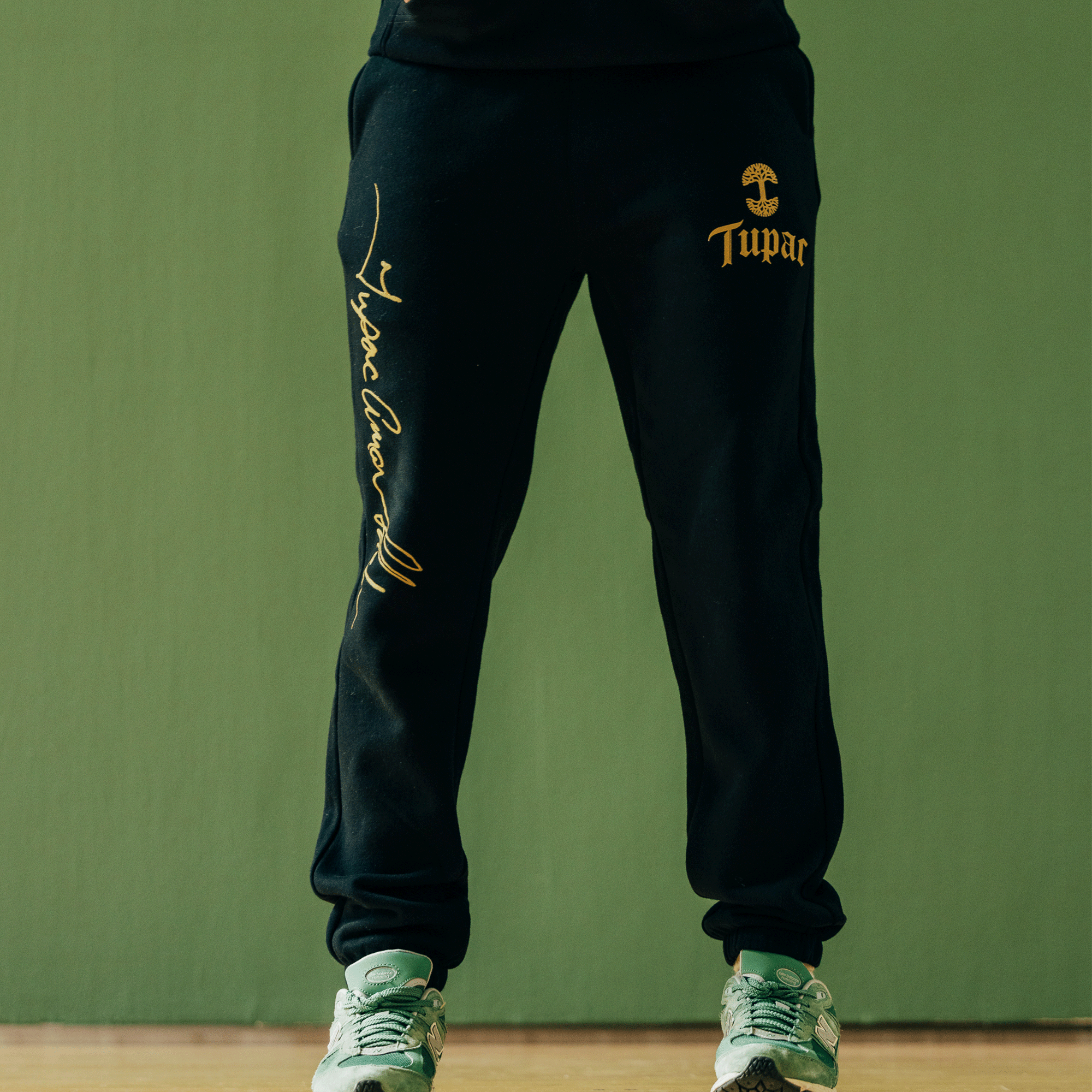 The person stands against a green backdrop, showcasing the "Bless Jogger" by Oaklandish, featuring "Tupac" in yellow script on the left leg. Sporting gray and green sneakers, the look captures the essence of Oaklandish x Tupac, highlighting the cultural impact of this unique collaboration on style.