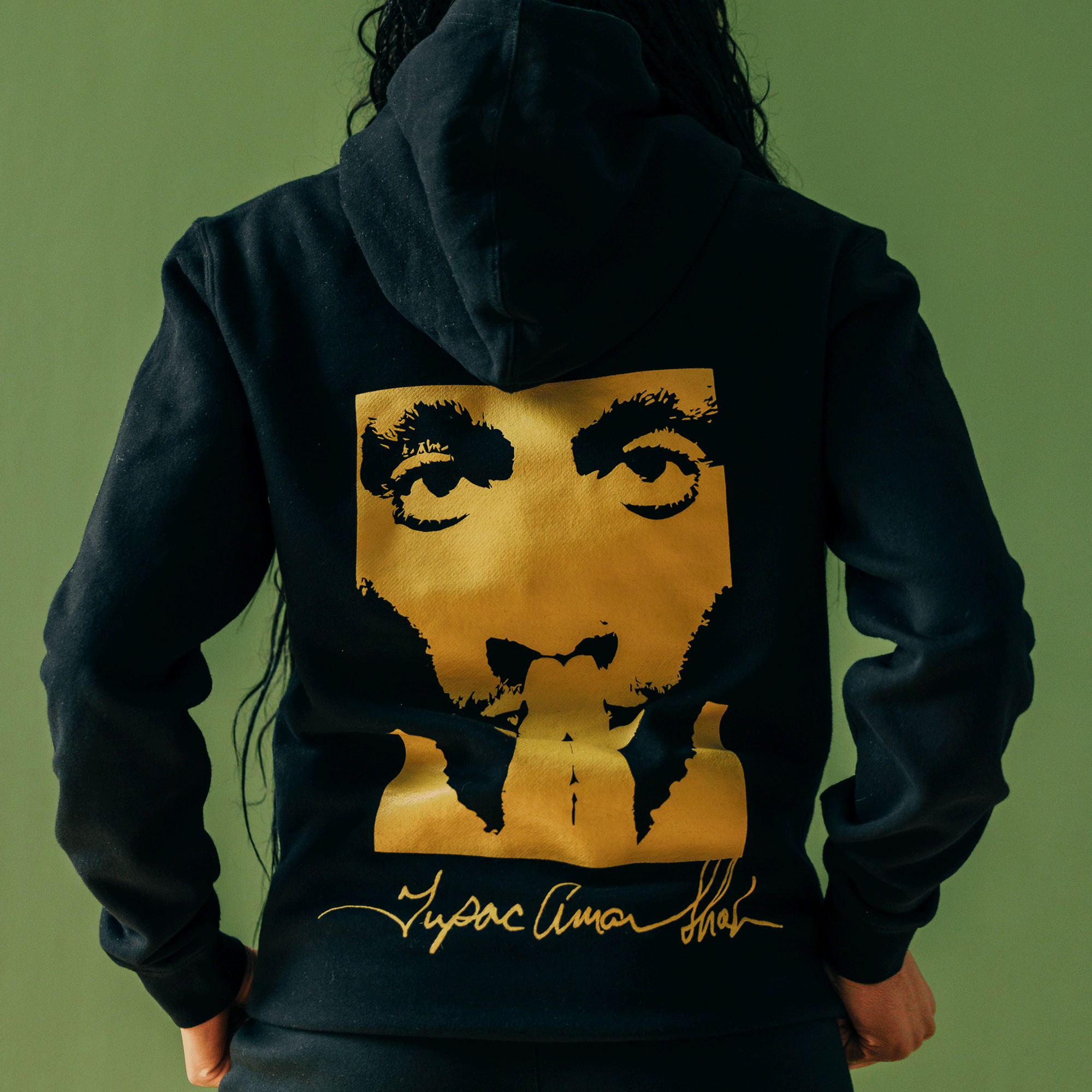 A person with long hair is wearing the Bless Hoodie from the Tupac collaboration by Oaklandish. This black hoodie features a striking gold graphic of a face with eyes and fingers over the lips alongside a gold cursive signature. The muted green background complements this piece from the iconic hip-hop collection.