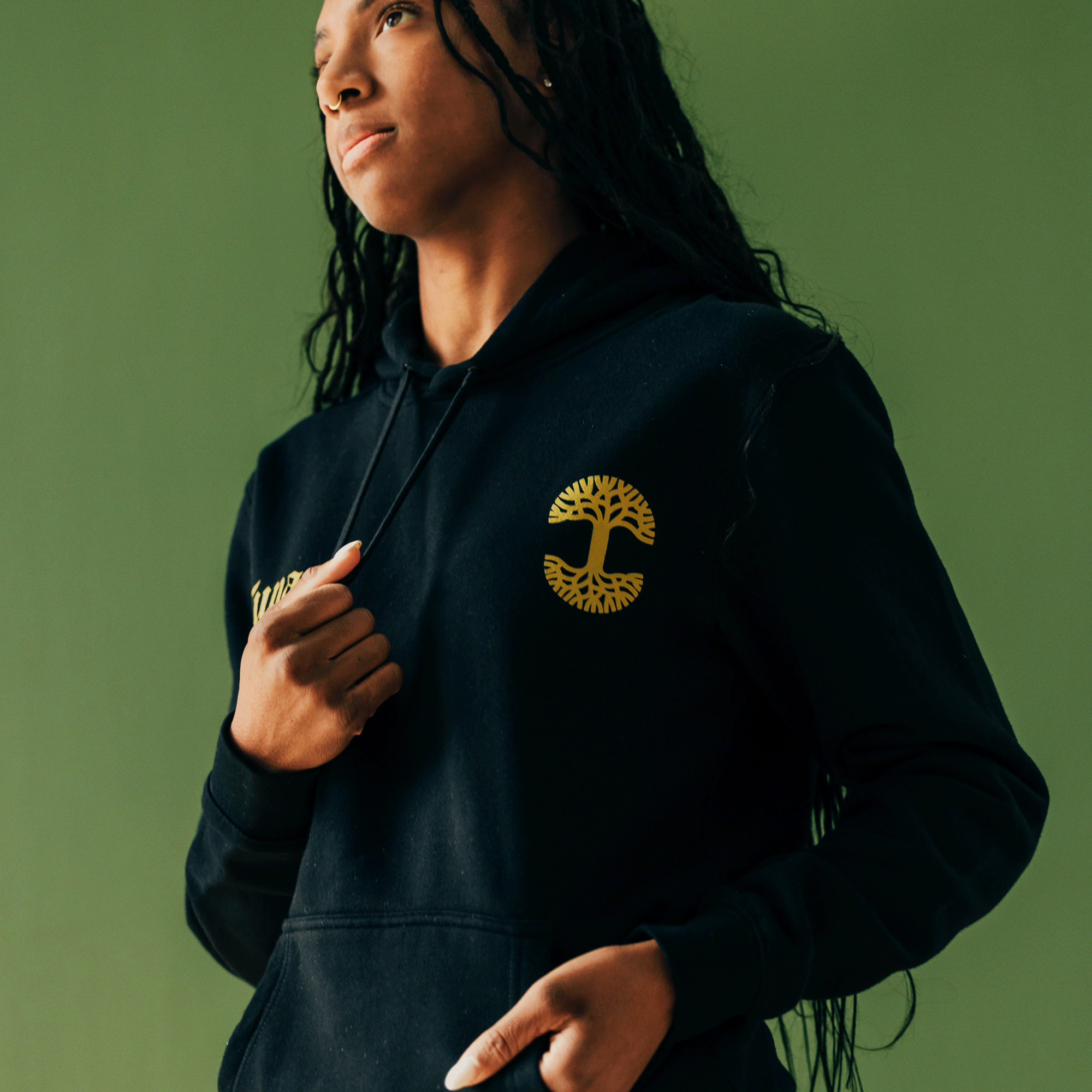 A person with long hair and a pensive expression stands against a green background, wearing the Bless Hoodie by Oaklandish. The black hoodie prominently displays the brand's stylized tree emblem on the chest. Their hand grasps the drawstring, and the soft lighting adds to the calm atmosphere of this piece from the hip-hop collection.