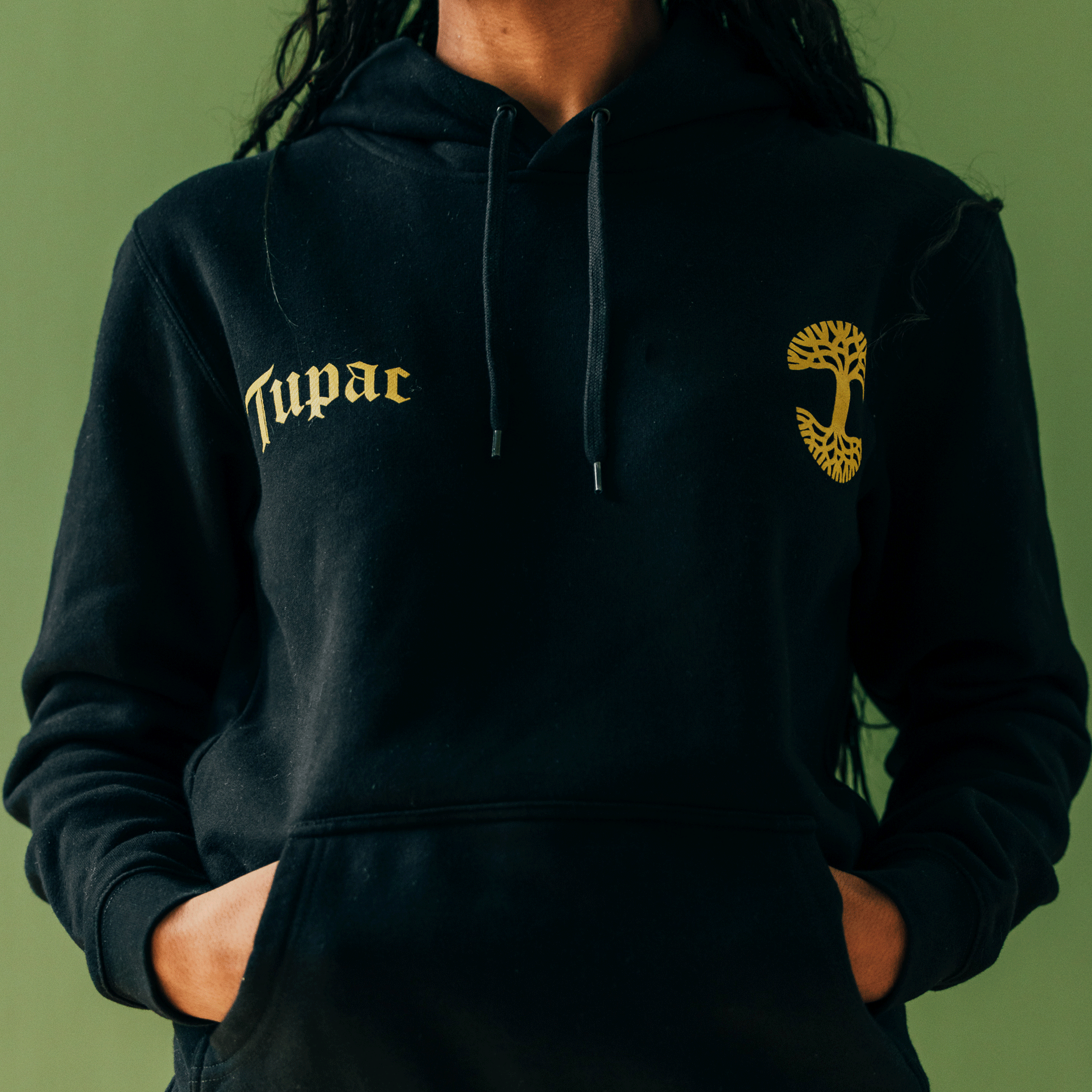 A person dressed in the Bless Hoodie by Oaklandish is standing against a green backdrop. This black hoodie features yellow Cyrillic text on the left chest and a yellow tree emblem on the right. Their face is hidden as their hands rest in the pockets, with long dark hair flowing down.