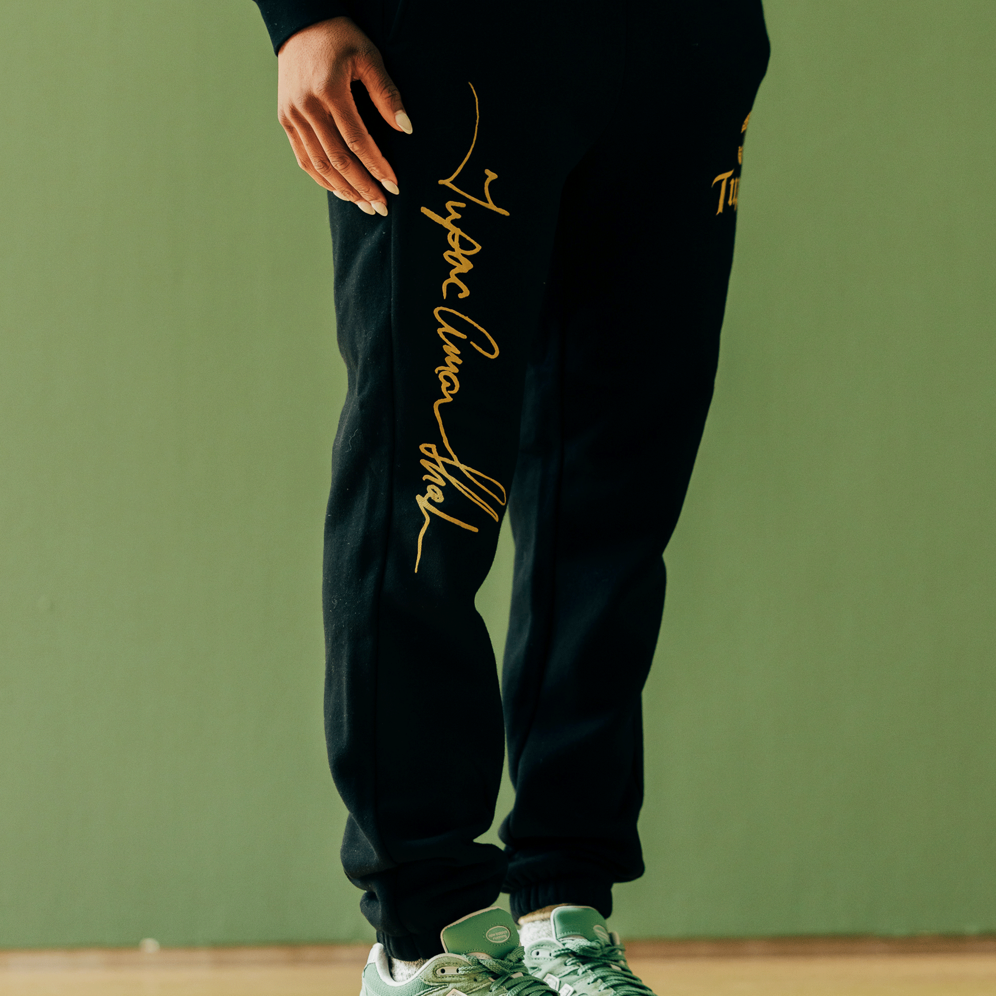 A person is wearing the Bless Jogger by Oaklandish, featuring gold cursive text on the side, standing against a green wall and showcasing their Tupac-inspired style. Only the lower half of their body is visible, stylishly paired with green and white sneakers. The text reads "More Than An Athlete," highlighting its cultural impact.