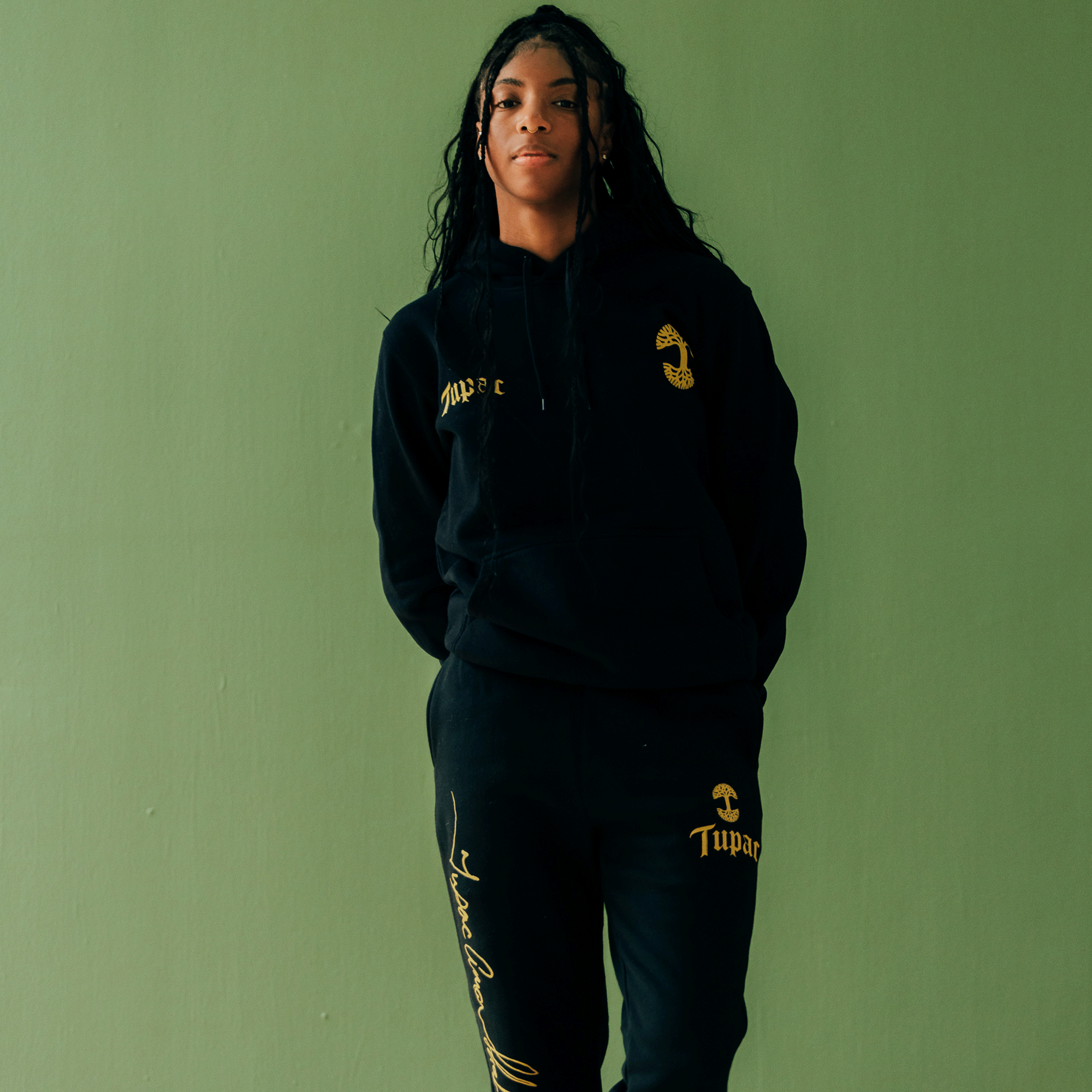 Amidst a green backdrop, a person with long, wavy hair showcases the Oaklandish Bless Hoodie in black paired with matching sweatpants featuring gold accents. This hip-hop collection honors iconic style as they stand confidently, hands behind their back, gazing directly at the camera.