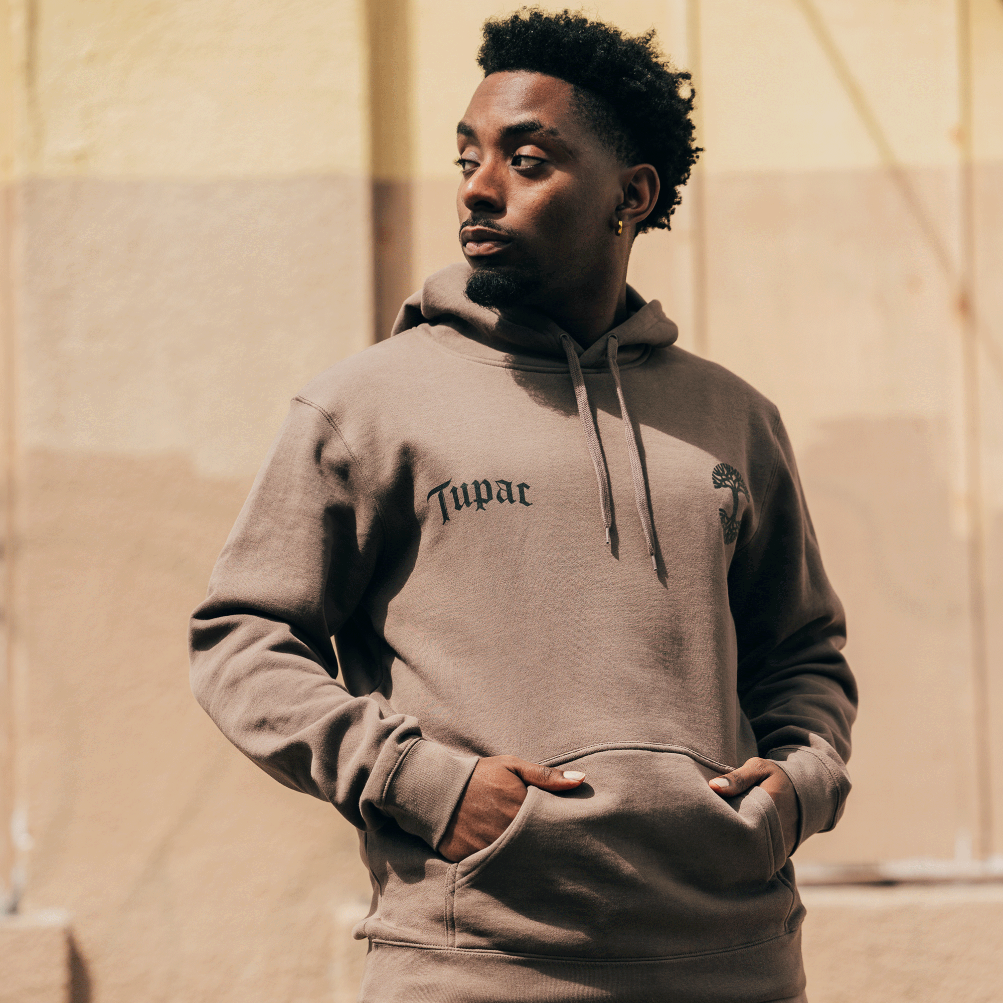 A man stands in front of a beige and yellow wall, wearing the Oaklandish Bless Hoodie in brown with "Tupac" boldly printed across the chest. With his hands tucked in his pockets, he gazes thoughtfully to the side, as if this piece were part of an exclusive Oaklandish x Tupac collection, casting soft shadows on the wall.