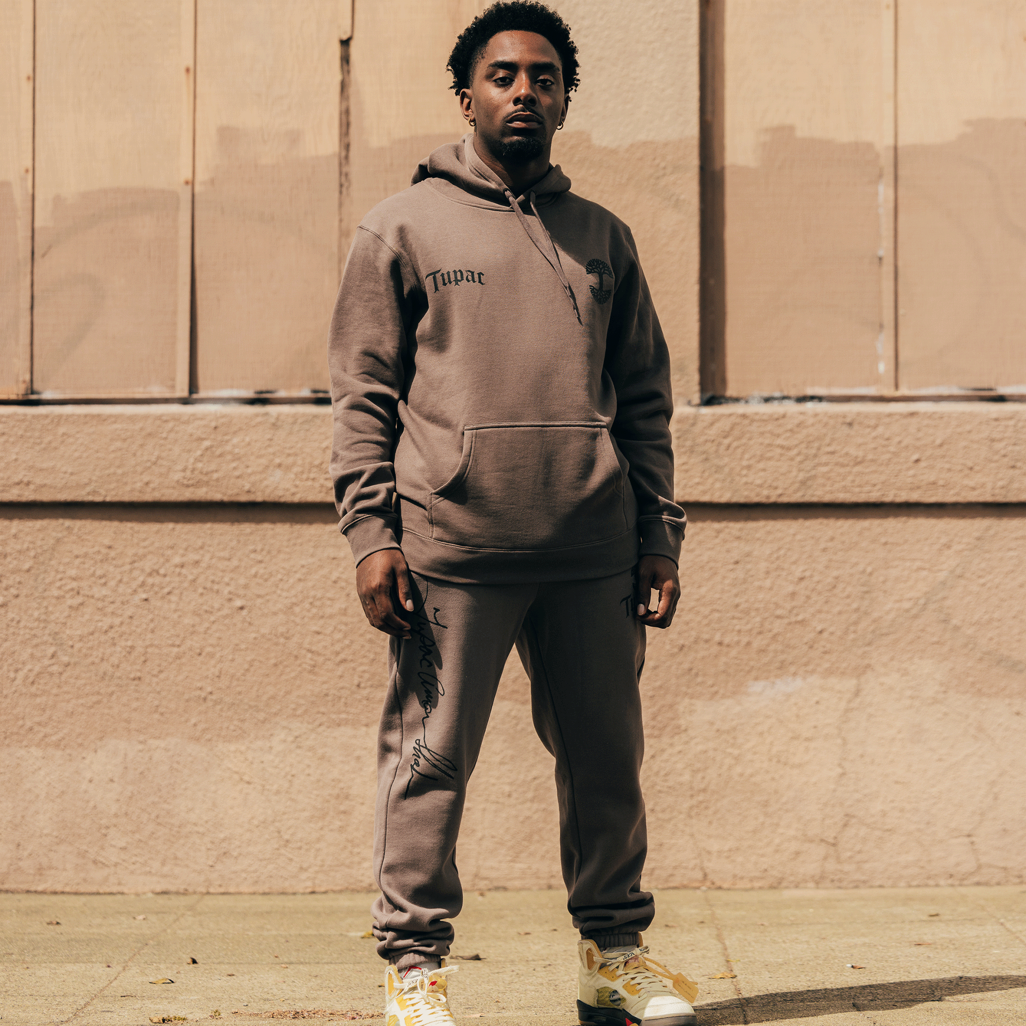 A man stands confidently outdoors against a textured, beige wall, wearing the brown Bless Jogger tracksuit from Oaklandish's Tupac collaboration hip-hop collection. With yellow sneakers and his hands in his hoodie pocket, he looks directly at the camera in a sunlit urban setting.