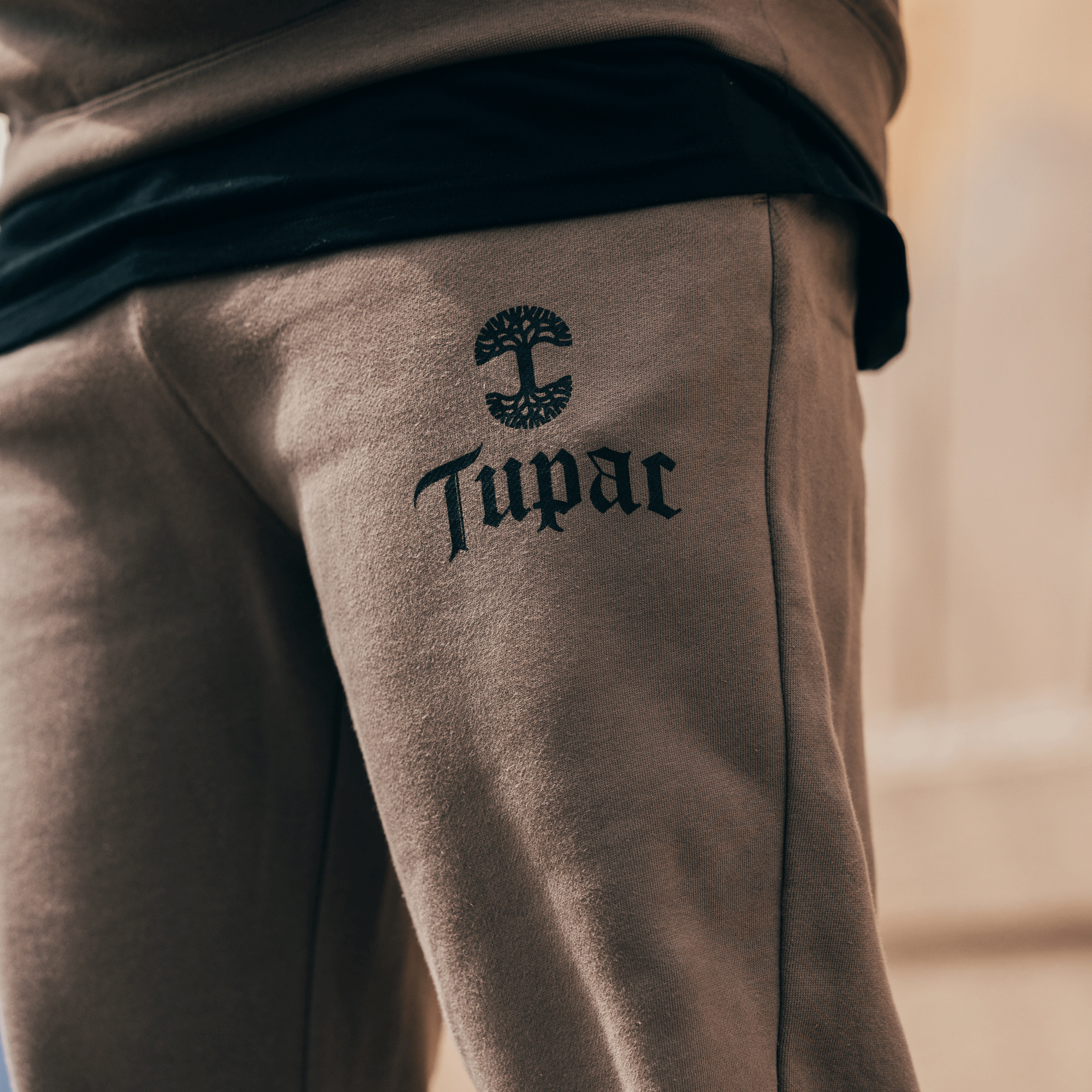 A close-up shot showcases someone wearing the Bless Jogger from Oaklandish, with "Tupac" and a tree symbol printed in black on the thigh. A black shirt is visible beneath the beige joggers, set against a softly blurred background that enhances the warm ambiance.