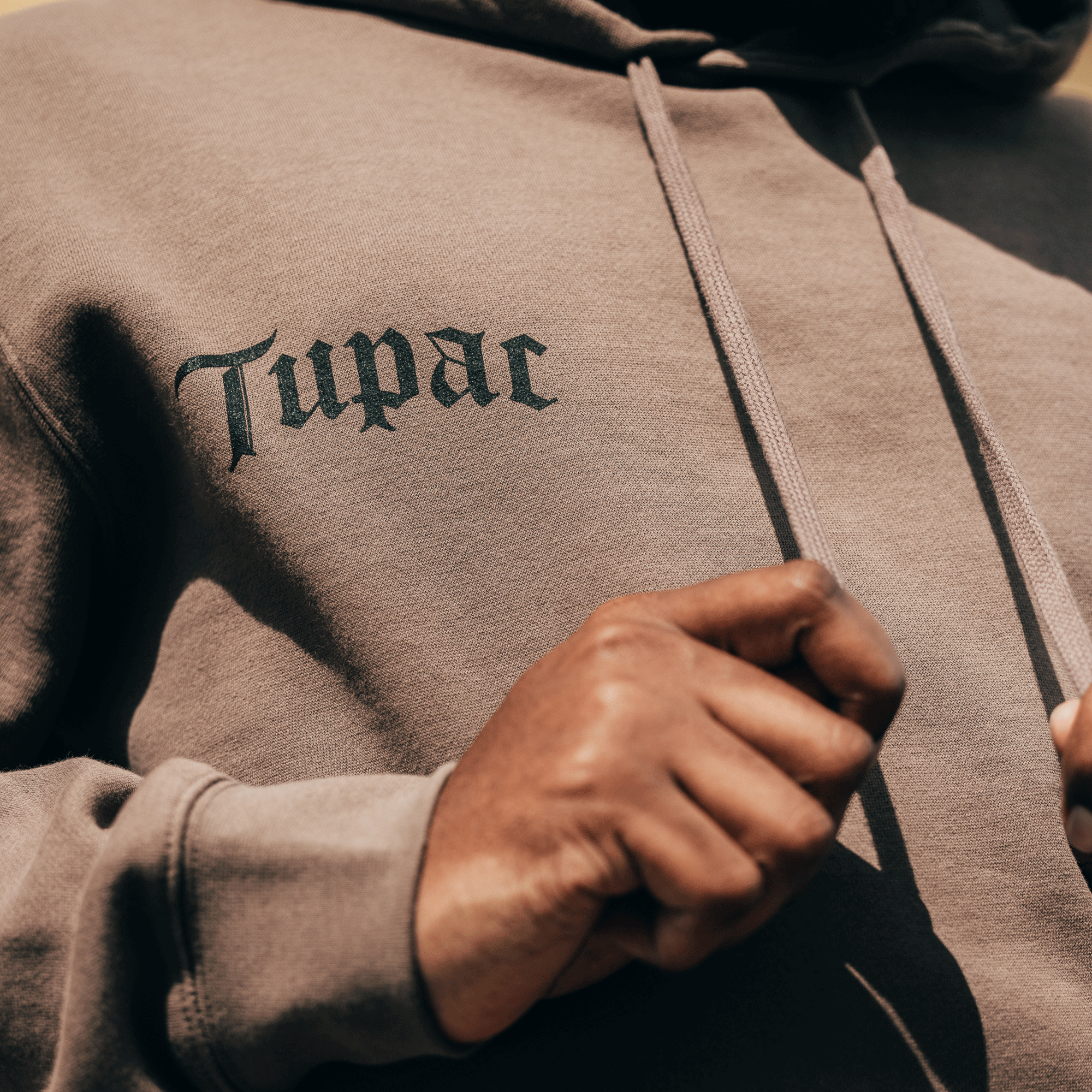 A person dressed in a taupe Bless Hoodie from Oaklandish highlights the "Tupac" embroidery in gothic script, representing the Oaklandish x Tupac partnership. Their hands gracefully hold the drawstrings as the softly blurred background emphasizes the lettering and fabric texture.