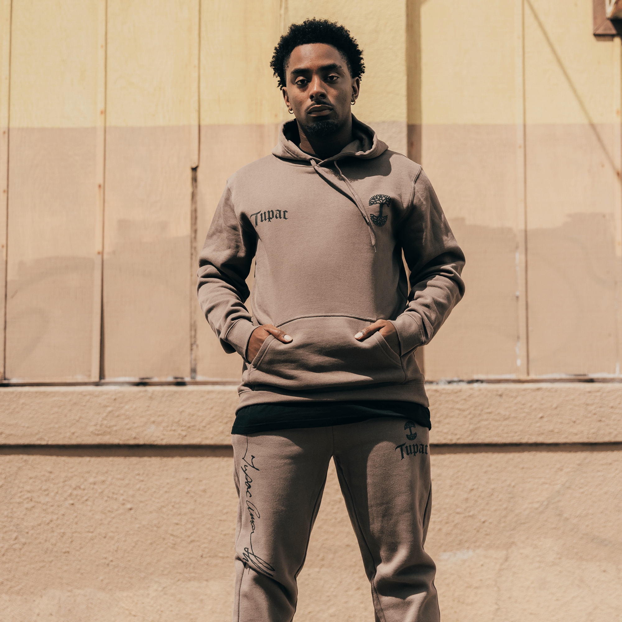 A person stands against a beige wall, dressed in an Oaklandish Bless Hoodie and matching brown sweatpants. Their hands rest in the hoodie pocket. The backdrop features a wooden panel with shadows and soft lighting, evoking the calm essence of Oakland hip-hop culture.