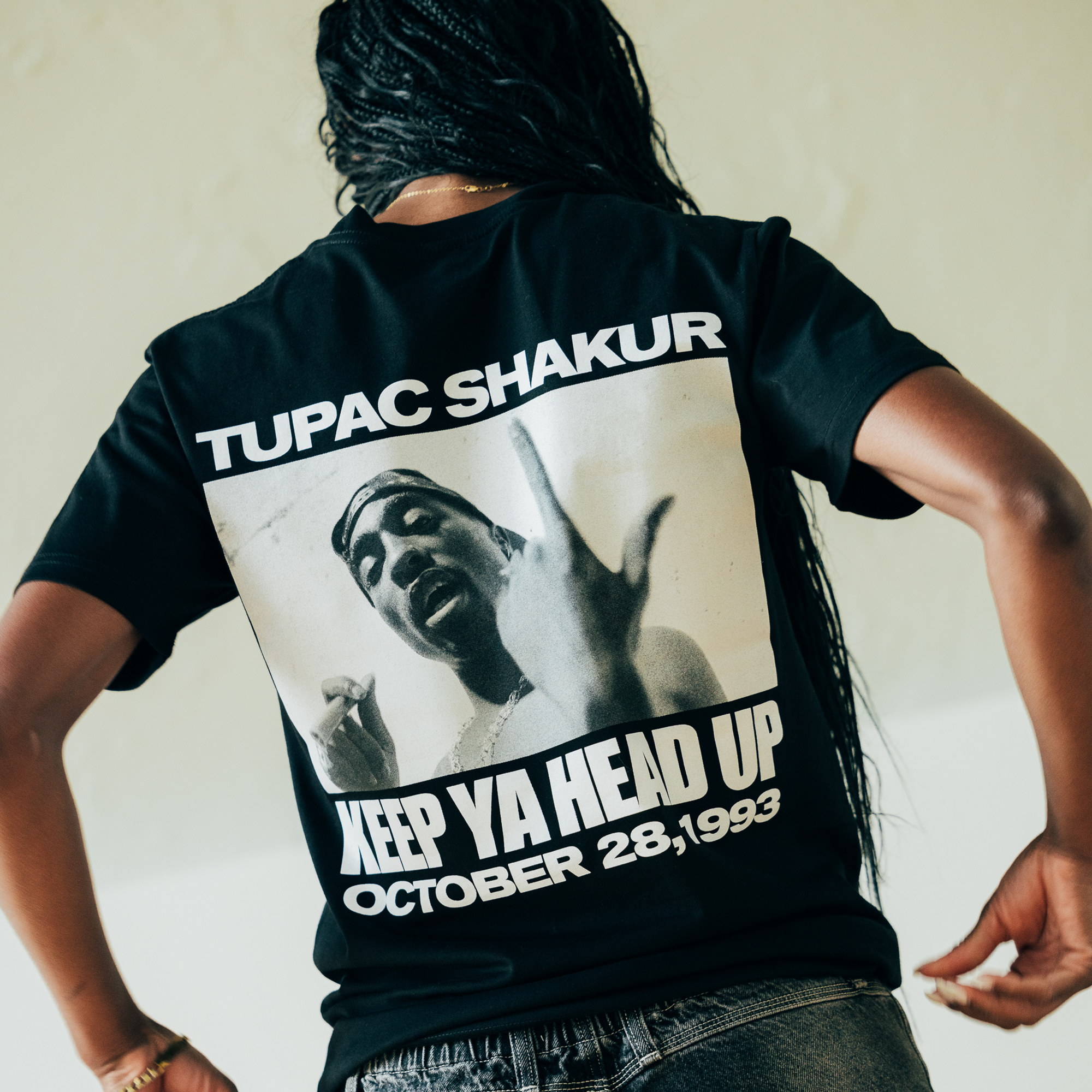 A person with long braided hair wears the Oaklandish Brighter Days Tee, which showcases a black-and-white image of Tupac Shakur gesturing with his hands. The text "KEEP YA HEAD UP" is accompanied by the date "OCTOBER 28, 1993." The individual faces away to highlight the back design of the T-shirt.