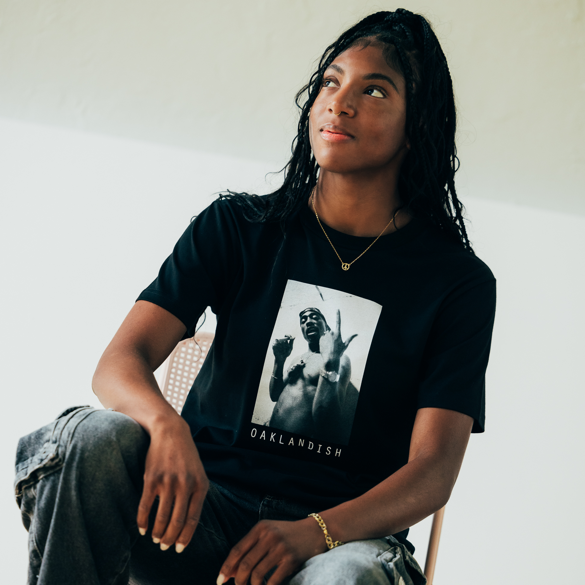 A person wearing a "Brighter Days Tee" from Oaklandish's hip-hop collection is seated on a chair. They have long, braided hair reminiscent of Tupac Shakur and are looking upward. A simple necklace and a bracelet adorn their left wrist. The background is plain, with soft lighting creating a calm atmosphere.