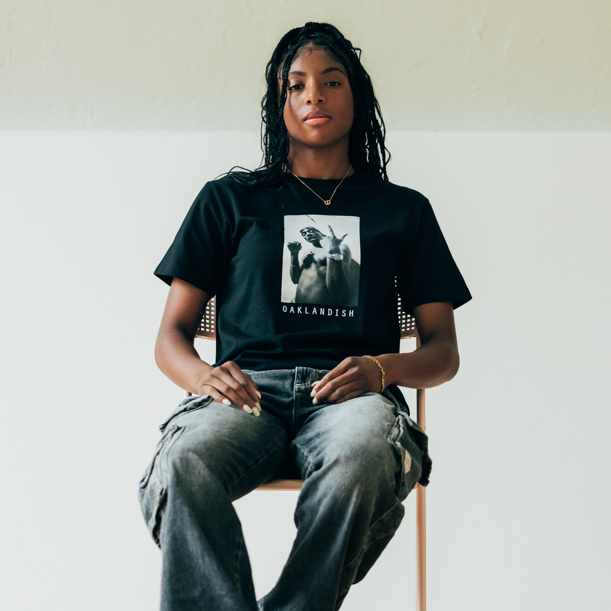 A person with long braided hair sits on a chair against a two-tone wall, capturing the essence of the Oaklandish style. They sport the Brighter Days Tee from Oaklandish, along with gray cargo pants. Their calm expression is highlighted by a watch and necklace.