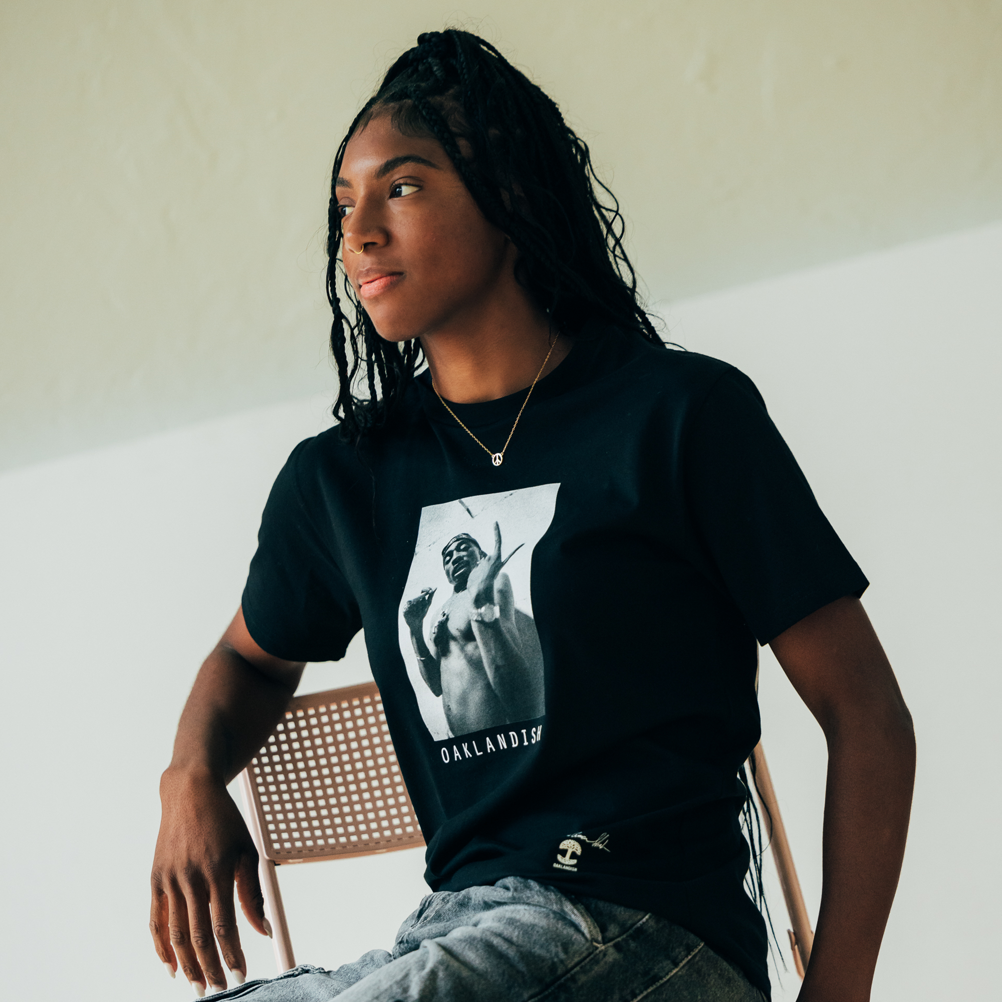 A person with long hair sits on a chair, wearing the Brighter Days Tee from Oaklandish's hip-hop collection, showcasing a graphic of Tupac Shakur in a dynamic pose. The individual is also dressed in light-colored jeans and has a thoughtful expression against a simple, light-colored wall.