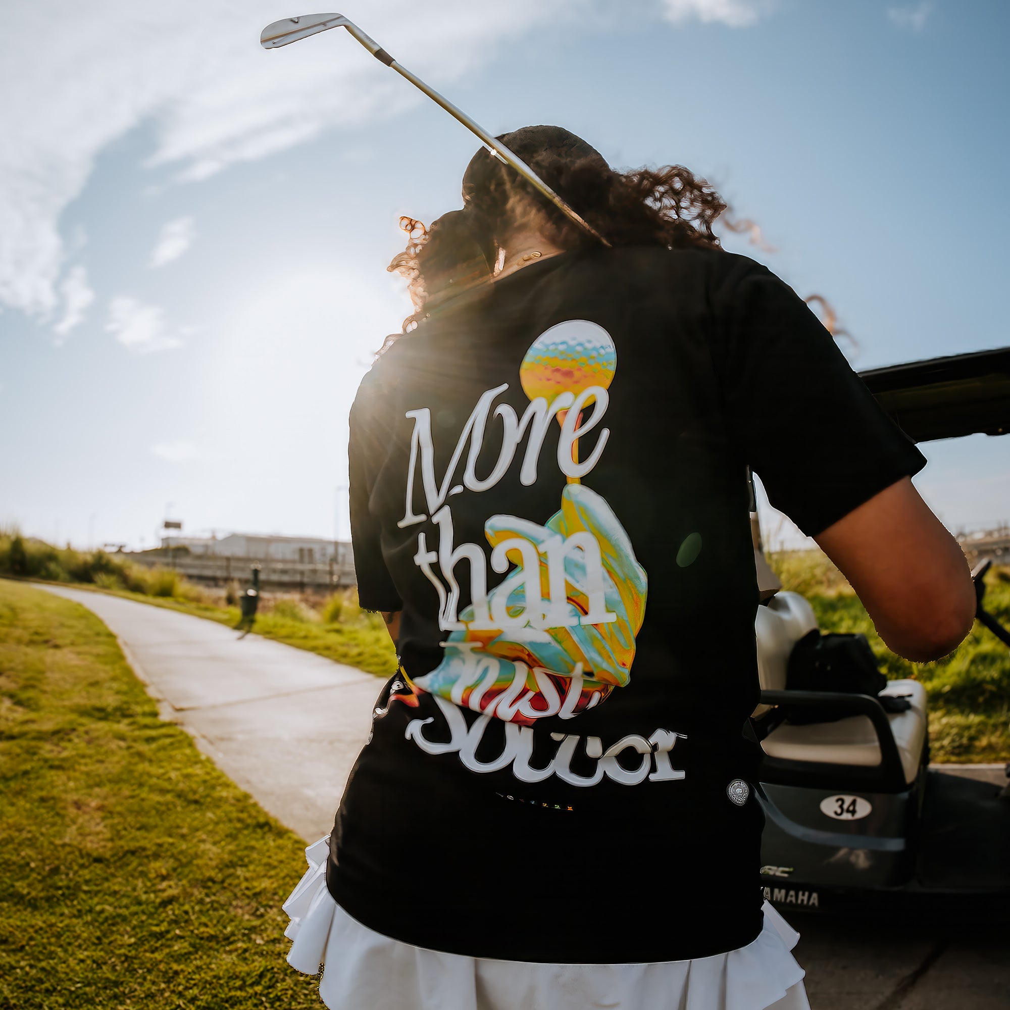 A person is standing on a grassy pathway near a golf cart, holding a golf club over their shoulder. They are wearing an Oakland Roots SC Classic fit t-shirt with colorful text that reads "More Than Soccer Tee." The sun shines brightly in the background, creating a vibrant and energetic atmosphere.