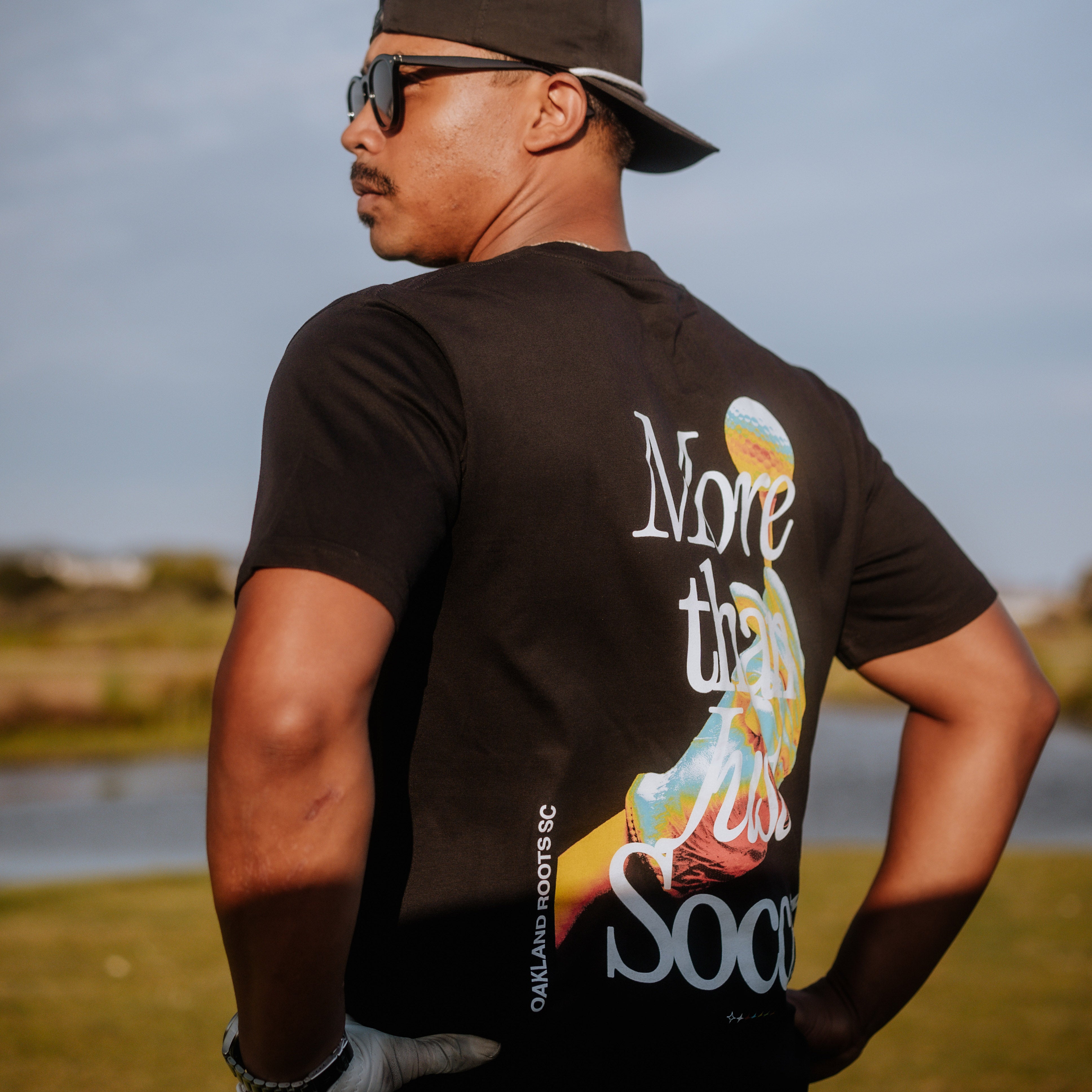 A man wearing an Oakland Roots SC "More Than Soccer Tee" with a graphic of a soccer player on the back stands outdoors. He sports an Oaklandish Classic fit t-shirt, a backwards baseball cap, sunglasses, and white gloves. The background features a grassy area and a body of water under a clear sky.