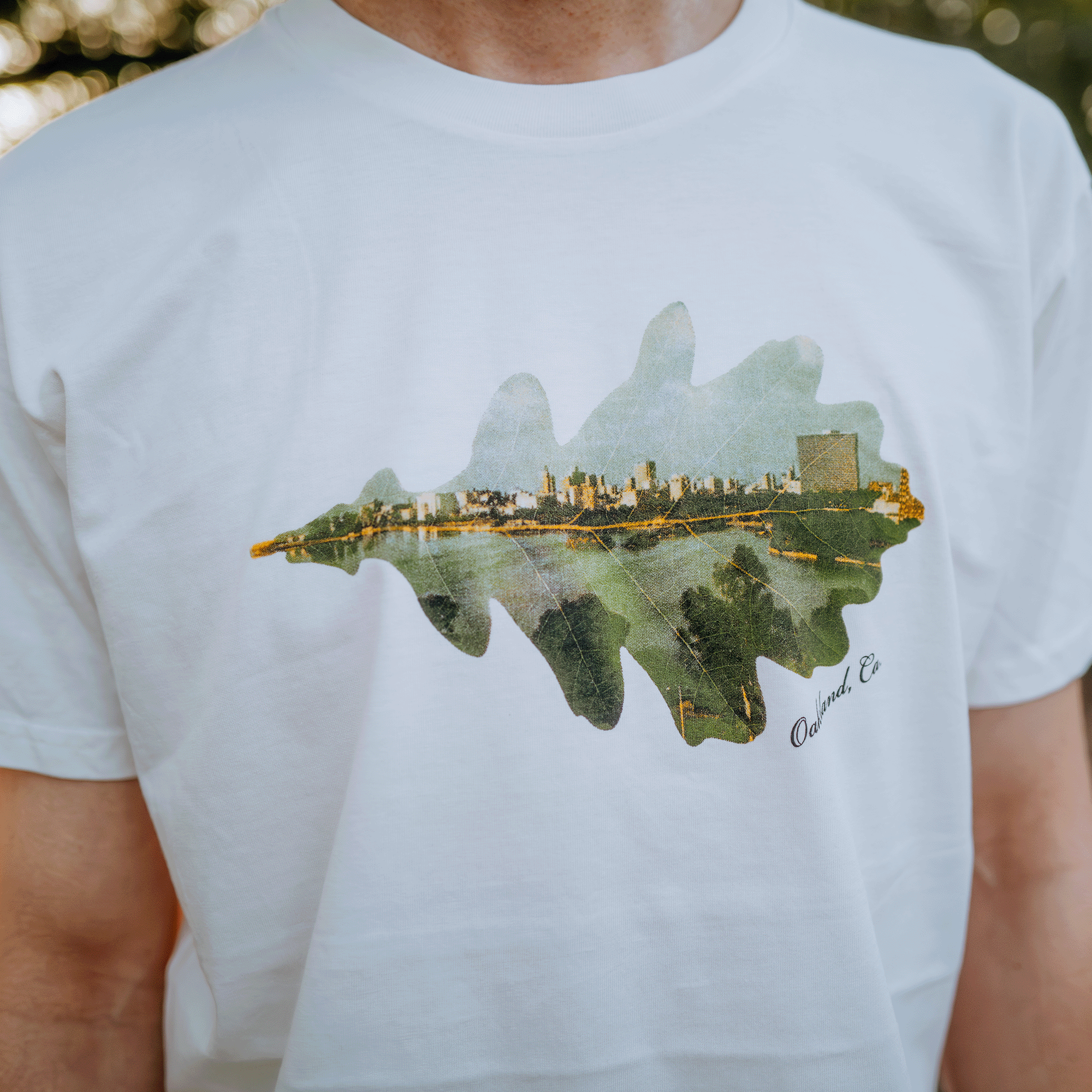 A person wearing the Double Exposure Tee by Oaklandish, a classic-fit, 100% cotton shirt that features a city's skyline fused with a large green leaf shape. This design elegantly combines urban and natural elements. The word "Oakland" along with "CA" is scripted at the edge of the leaf against blurred greenery.