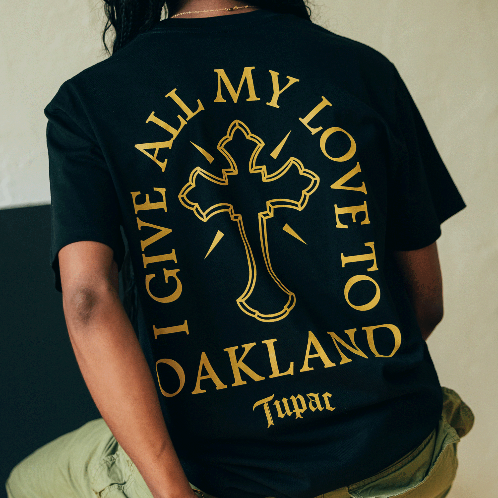 A person wears the Oaklandish Double Cross Tee, featuring a black T-shirt with a bold gold-colored design on the back. The design captures an Oaklandish spirit with a stylized cross surrounded by rays and the text "I GIVE ALL MY LOVE TO OAKLAND Tupac." The individual is seated, highlighting the shirt's distinctive back design.