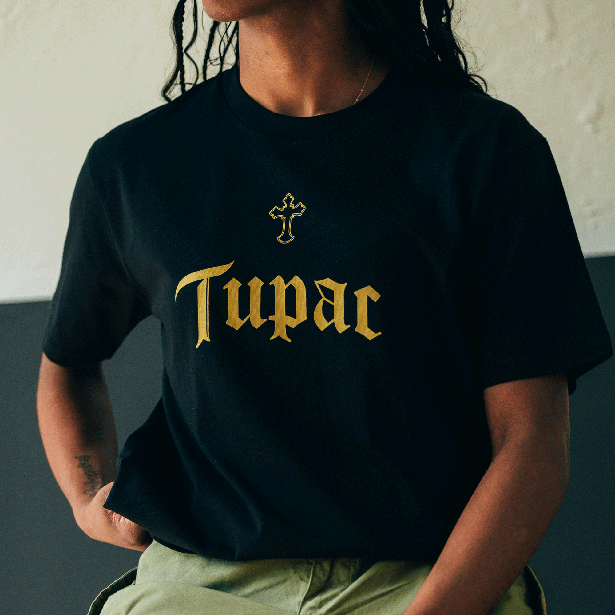 Wearing the Oaklandish Double Cross Tee, a person exudes a distinct hip-hop vibe. This black T-shirt features "Tupac" in gold Gothic lettering with a gold cross above the name. With long hair and one hand casually in a pocket, they sit against a neutral background, creating an effortlessly minimalistic look.