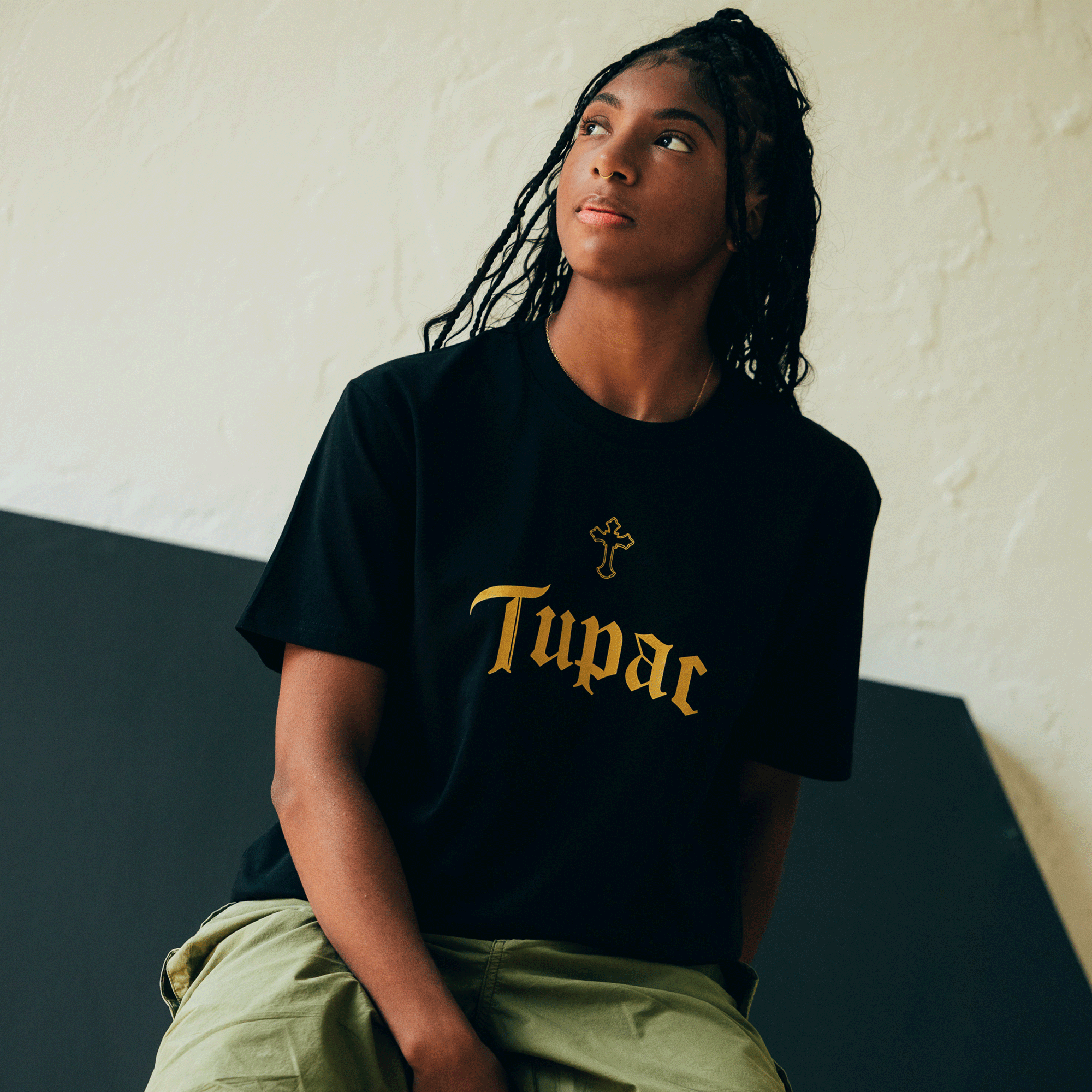 A person with long braided hair is sitting and gazing upward, exuding the essence of hip-hop culture. They are wearing a black "Double Cross Tee" by Oaklandish, which features a gold cross design and is paired with green pants. The backdrop is a straightforward two-tone wall in beige and black, enhancing the Oaklandish atmosphere of the scene.