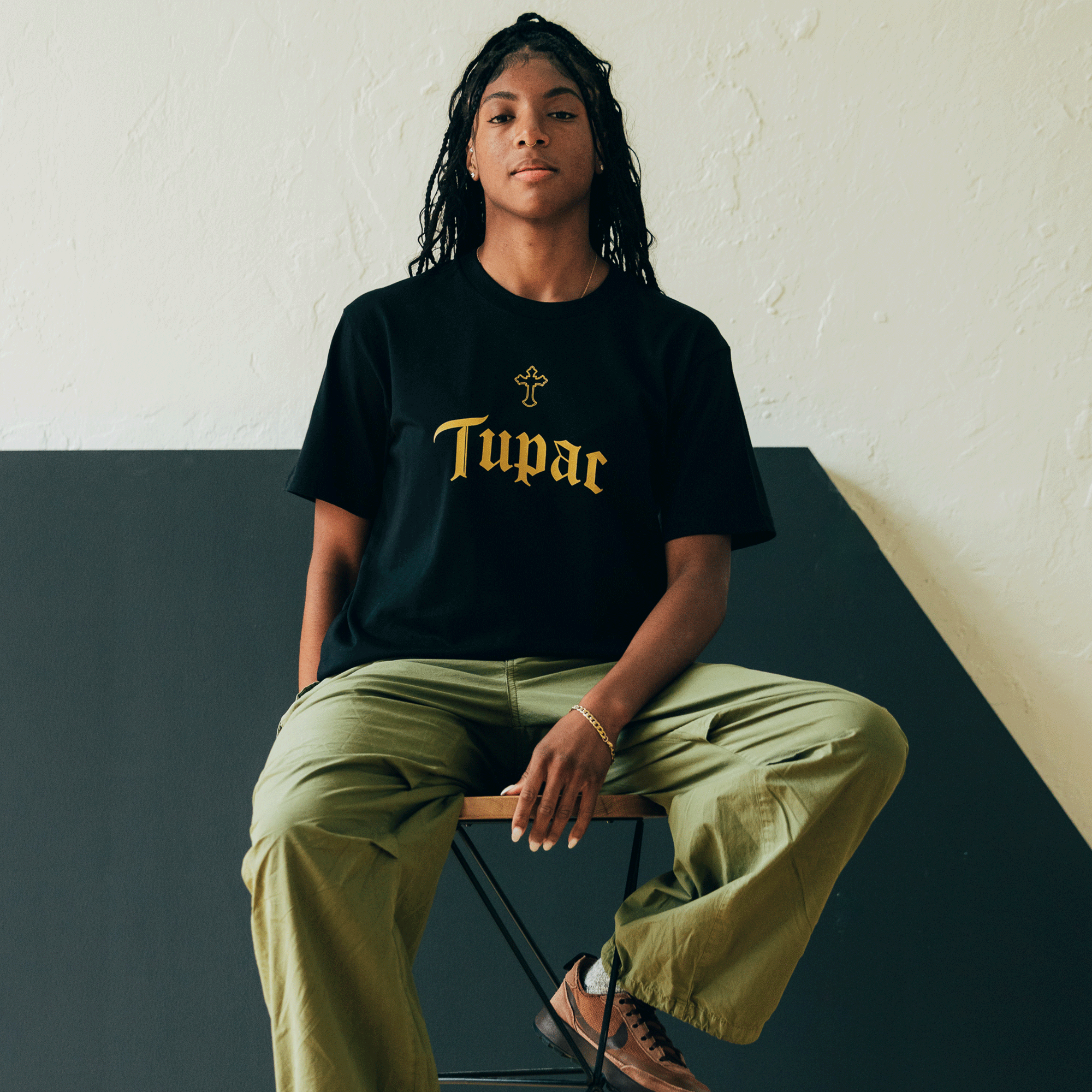 A person with long hair sits on a stool against a two-toned wall, wearing the Oaklandish Double Cross Tee featuring "Tupac" and a cross symbol in gold lettering. They sport olive-green pants and brown shoes, exuding hip-hop vibes.