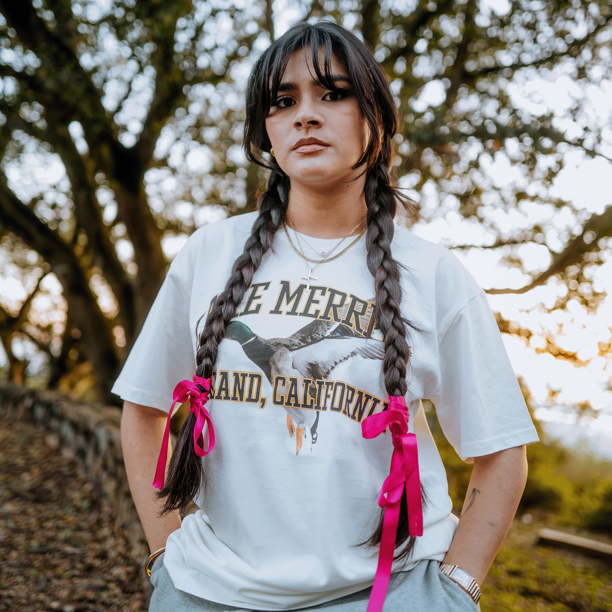 A woman with long braided hair tied with pink ribbons stands outdoors, wearing the Duck School Tee by Oaklandish. This classic fit, 100% cotton white t-shirt showcases a graphic with partially visible text. The background features trees basking in the warm glow of sunset.