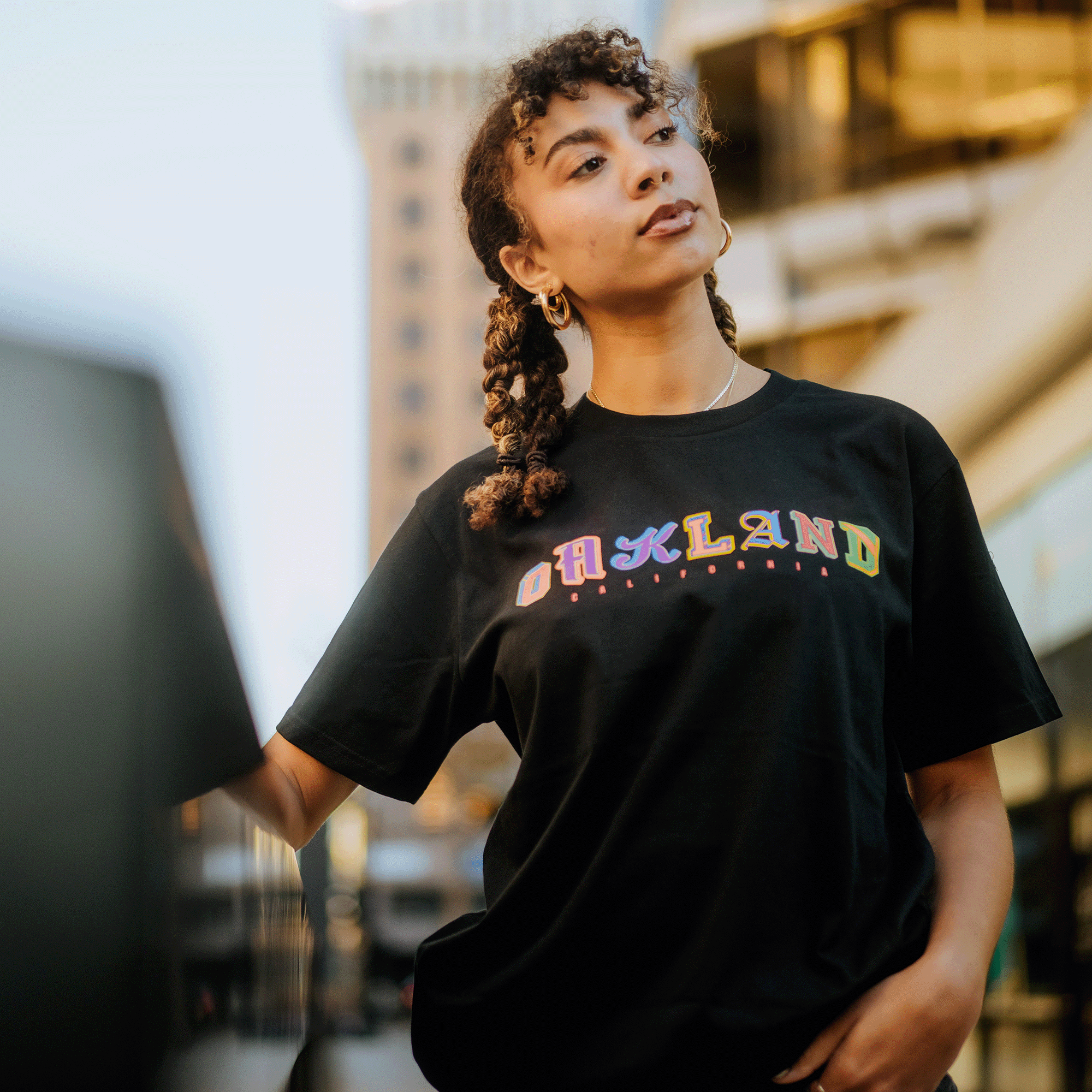 A person with curly hair, wearing gold hoop earrings and the First Team Tee—a black classic fit T-shirt made from heavy-weight cotton featuring vibrant "OAKLAND" lettering by Oaklandish—stands outdoors. The background features a blurred urban setting with a tall building and lights. The person gazes to the side, holding onto a nearby structure.