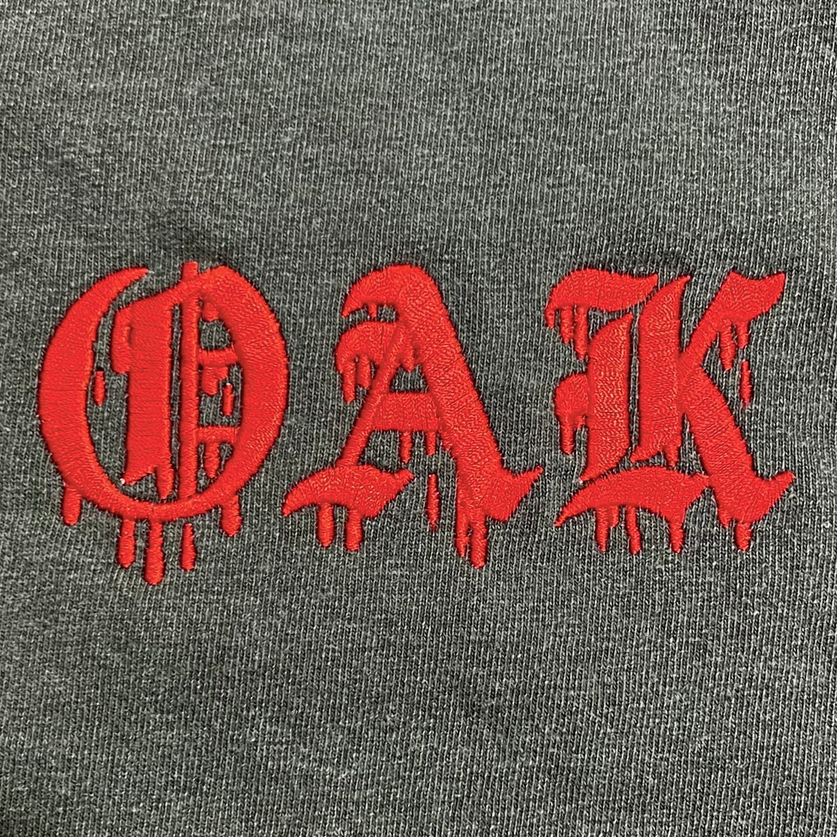 The Bleed Tee by Oaklandish features the OAK logo in a red Gothic font with blood-like dripping effects, set against a dark gray background. This t-shirt offers a classic fit and is crafted from heavyweight cotton, combining style and durability effortlessly.