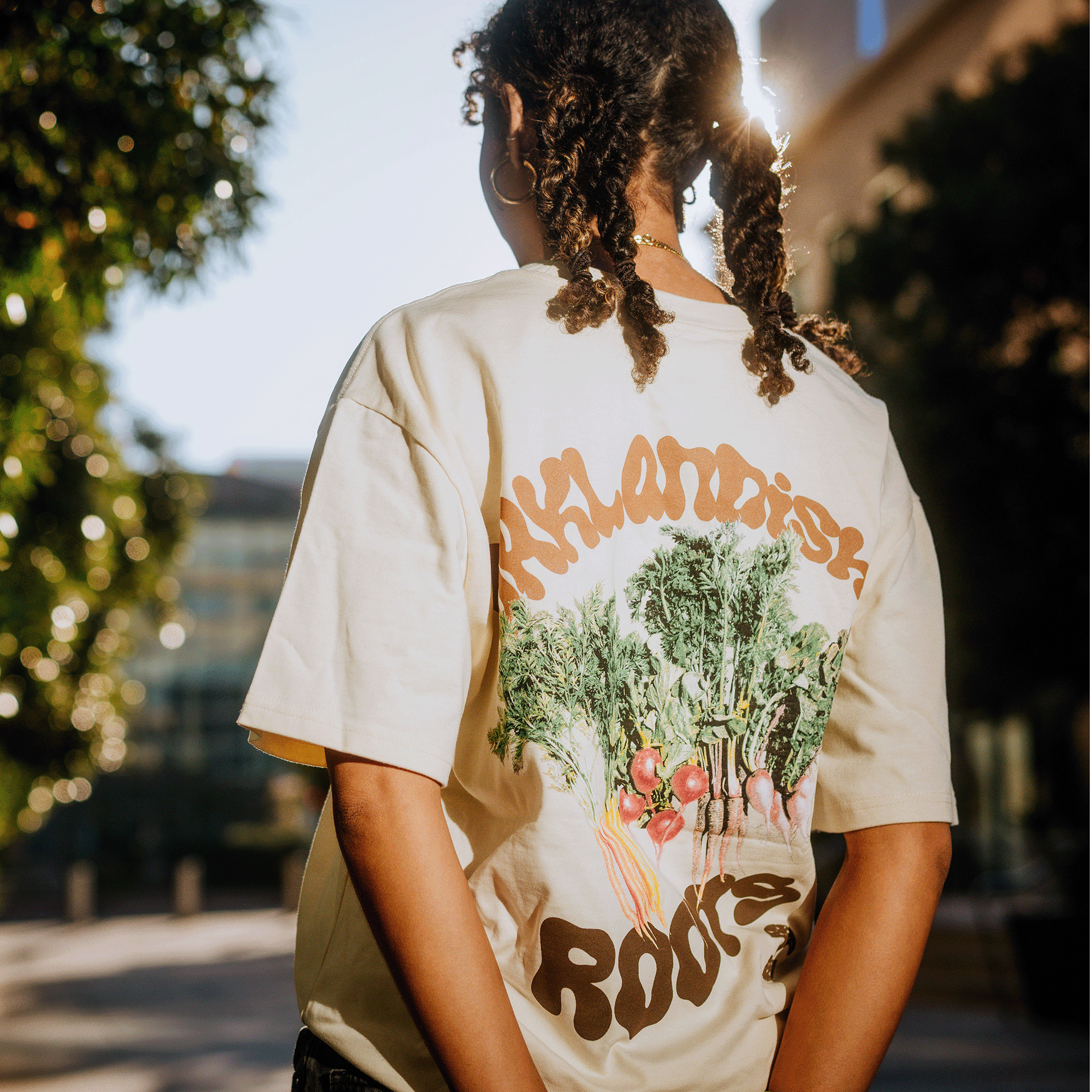 Standing outdoors with braided hair, the person faces away while wearing the Grow Your Roots Heavy Tee by Oaklandish. The boxy fit men's t-shirt, featuring colorful vegetables and "KALAMATA'S ROOTS" on the back, is made from heavyweight combed cotton. Sunlight filters through the trees, highlighting the fabric in a warm, natural setting.