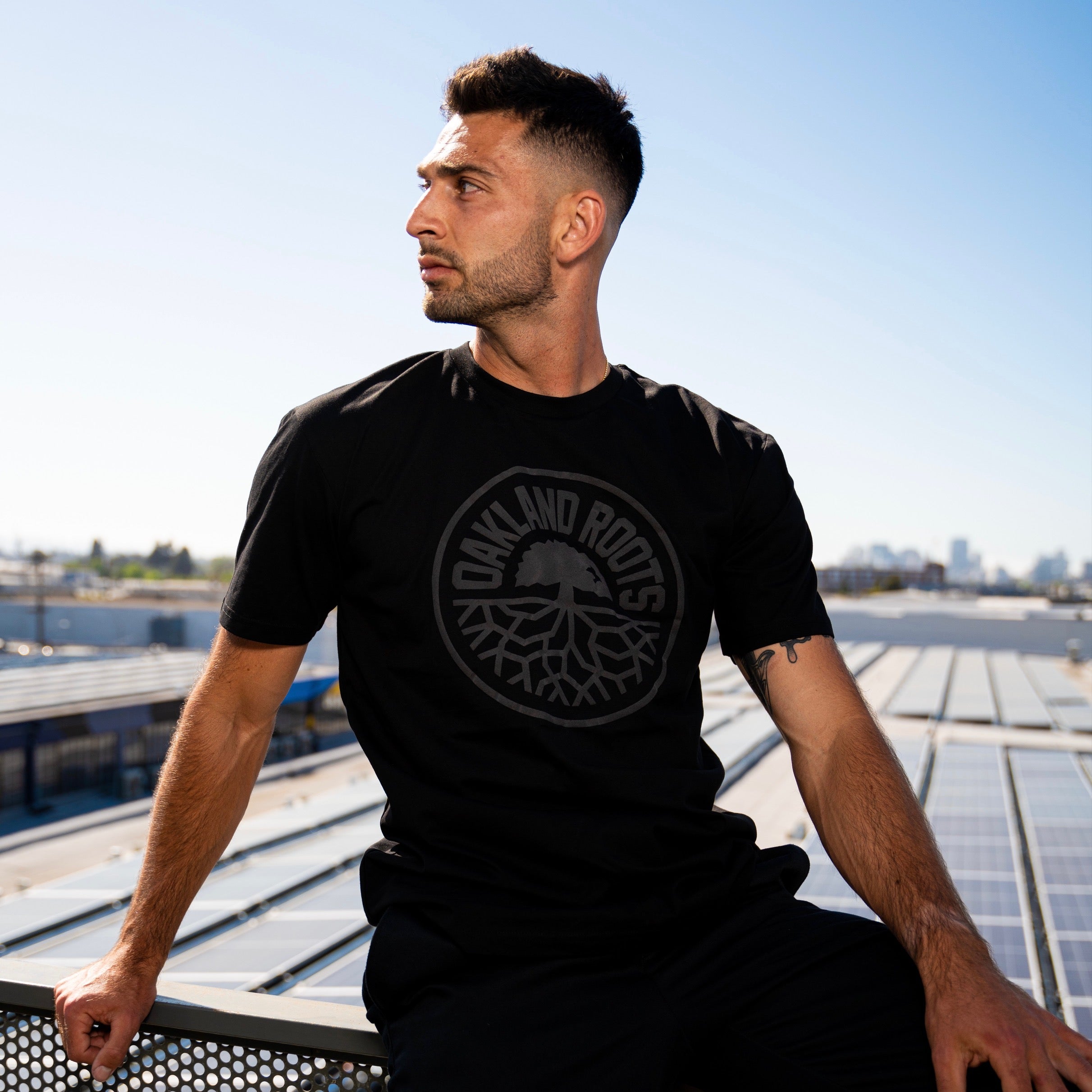 A man with short dark hair and a trimmed beard, wearing an "Oakland Roots SC Blackout Logo Tee," sits on a metal railing. He looks to the left with a thoughtful expression. The background features a rooftop with solar panels and a distant city skyline under a clear blue Oaklandish sky.