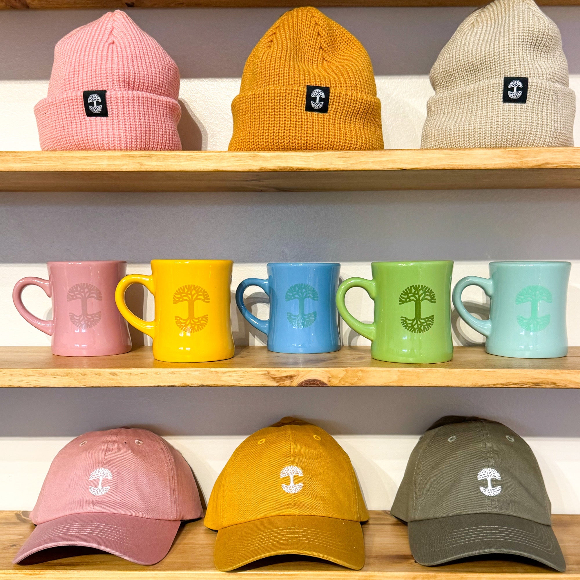 Shelves showcase a charming collection of Oaklandish drinkware: four ceramic Oaklandish Diner Mugs in pink, yellow, blue, and green on the middle shelf. The top shelf features three knitted beanies in pink, mustard, and beige. Below, you'll find three baseball caps in pink, yellow, and olive green. Every item proudly displays a tree logo.