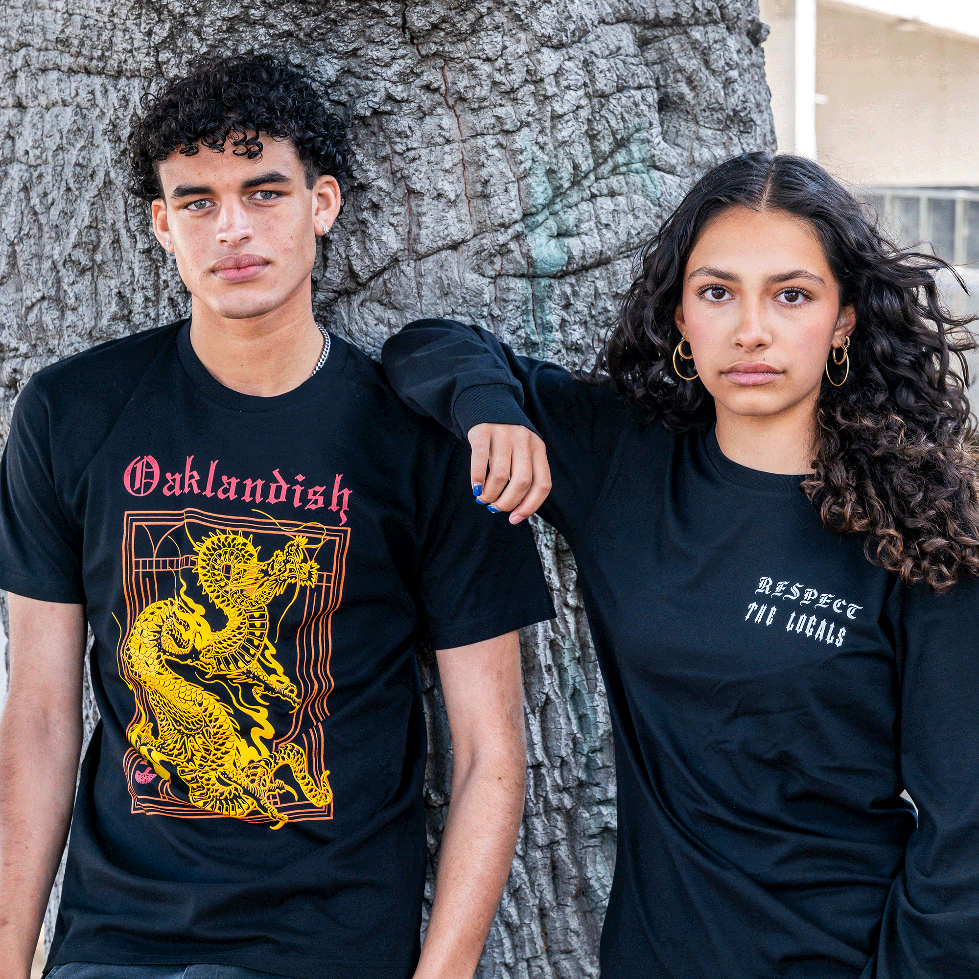 A young man and woman lean against a tree, both wearing classic fit black Prosperity Tees from Oaklandish. The man's shirt showcases a yellow dragon with the word "Oaklandish," while the woman's tee reads "RESPECT THE LOCALS." With curly hair, they gaze peacefully at the camera.