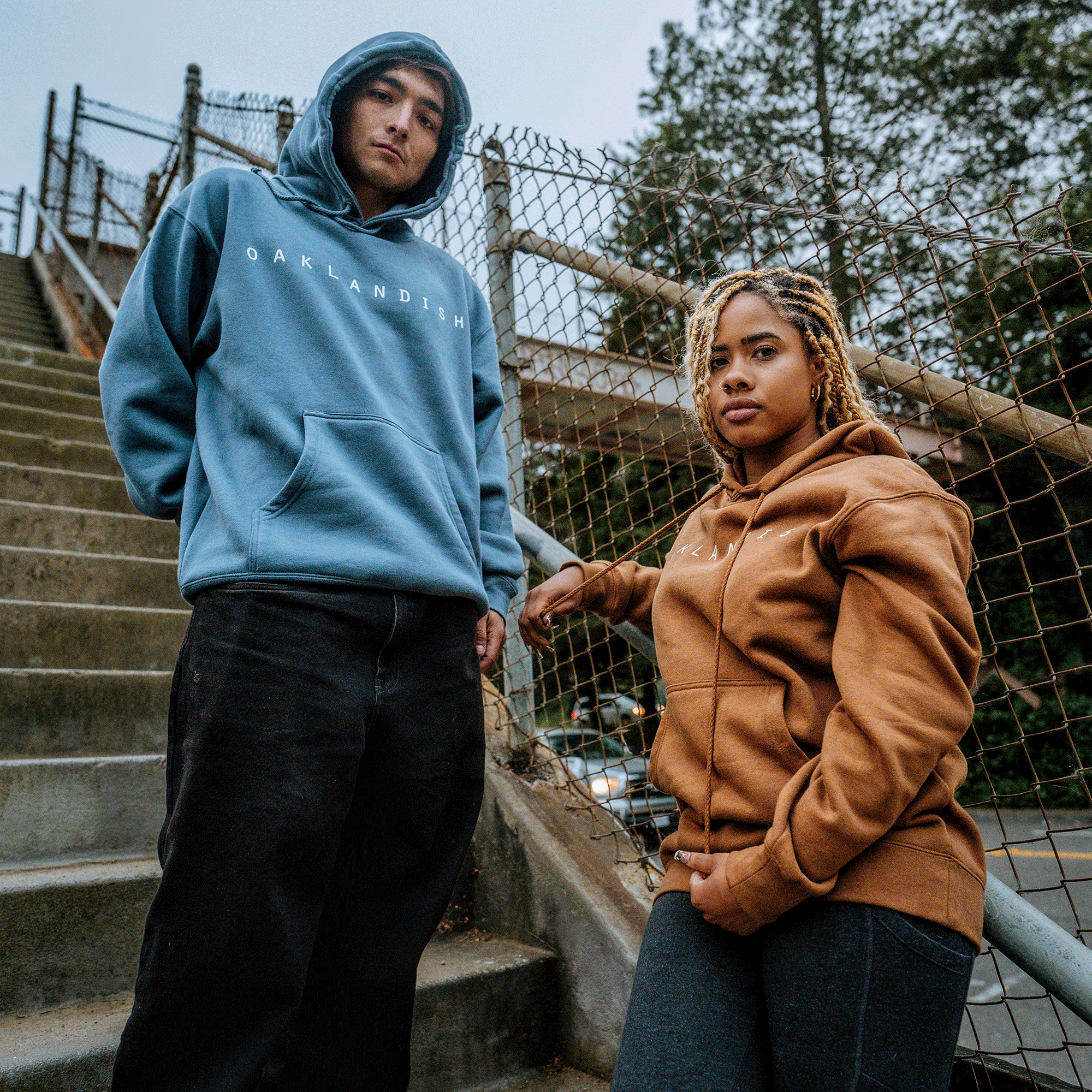 Two individuals stand confidently on a stairway near a chain-link fence. The person on the left wears an Oaklandish Classic Hoodie in storm blue, while the person on the right sports an Oaklandish Classic Hoodie in brown. Both hoodies proudly display "Oaklandish," showcasing their Oakland pride. Trees and a walkway are visible in the background, and their expressions are serious.