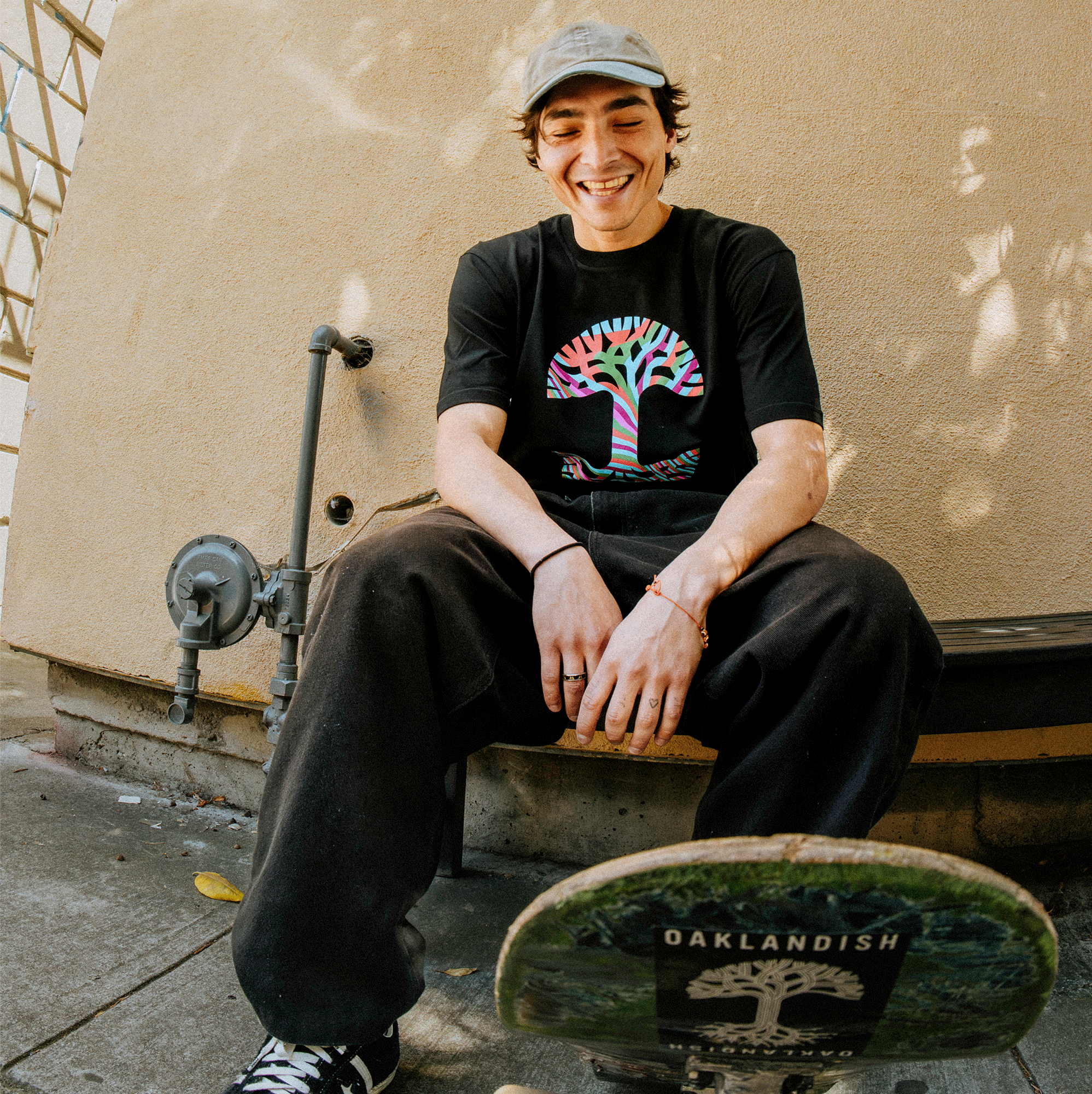 A smiling young man wearing a Stripe Tree Tee, a 100% cotton and classic fit t-shirt from Oaklandish featuring a colorful tree design, sits on a bench holding a skateboard that has "OAKLANDISH" written on it. He is in a sunny outdoor setting with a beige wall and some pipes next to him, showcasing the men's collection.