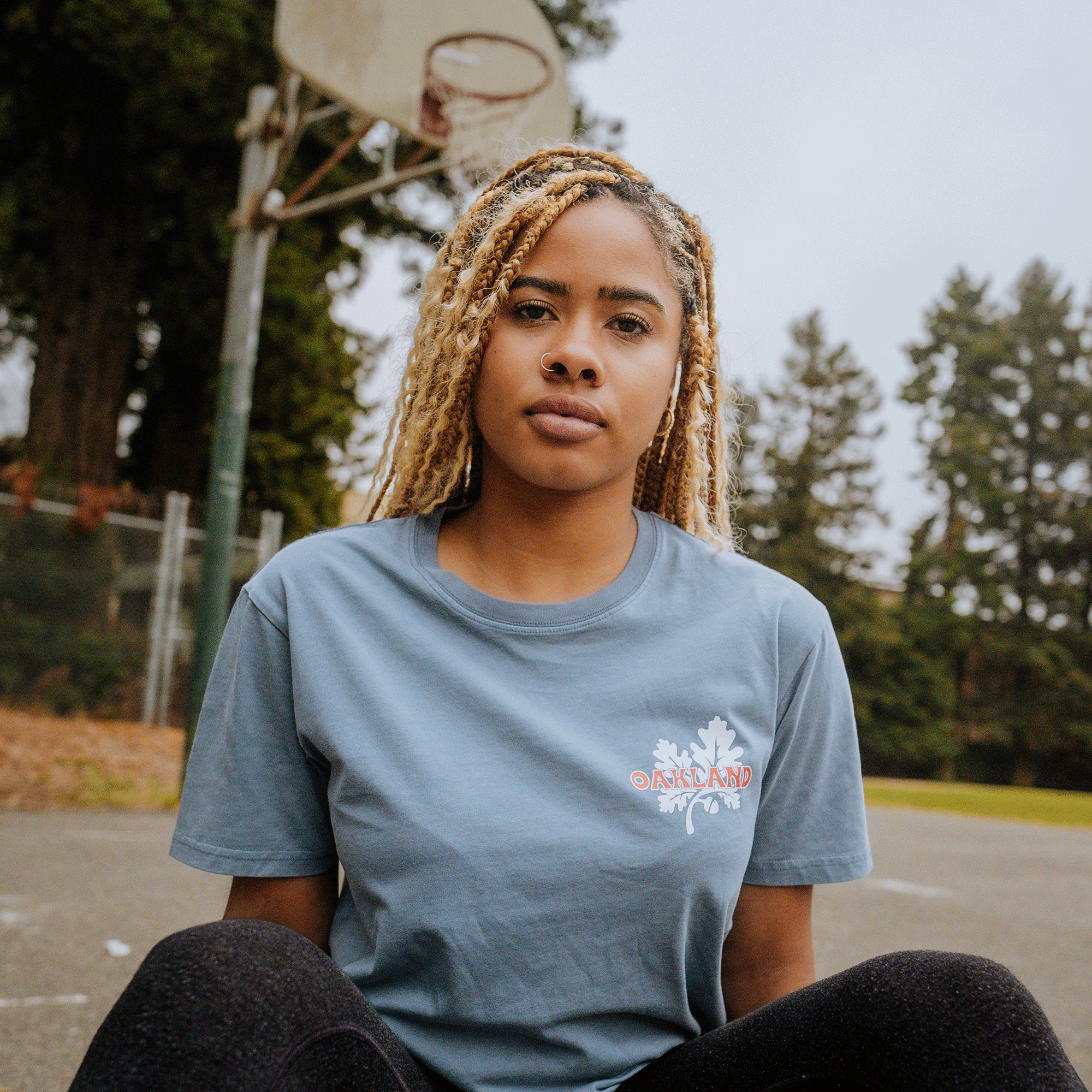 A woman with light brown braided hair is sitting on an outdoor basketball court, looking at the camera. She is wearing a classic fit "Grocery Run Tee" in light blue with the word "OAKLAND" on it and black pants, both by Oaklandish. The freshest basketball hoop and tall green trees are visible in the background. It is a cloudy day.