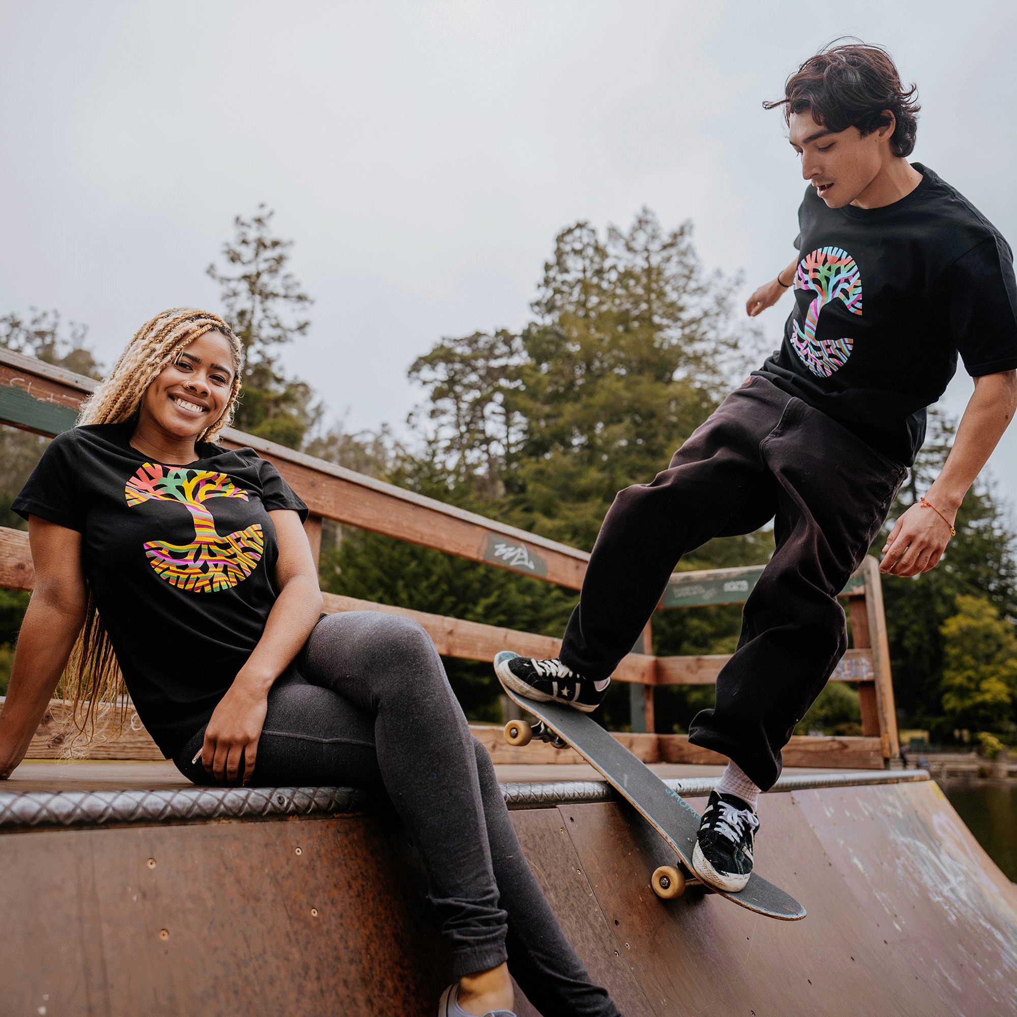 A smiling woman with braided blonde hair sits on a ramp, wearing a black classic fit Women's Stripe Tree Tee from Oaklandish and gray pants. Next to her, a young man with dark hair skateboards, wearing a matching T-shirt and dark pants. They are outdoors on a cloudy day, with trees in the background.