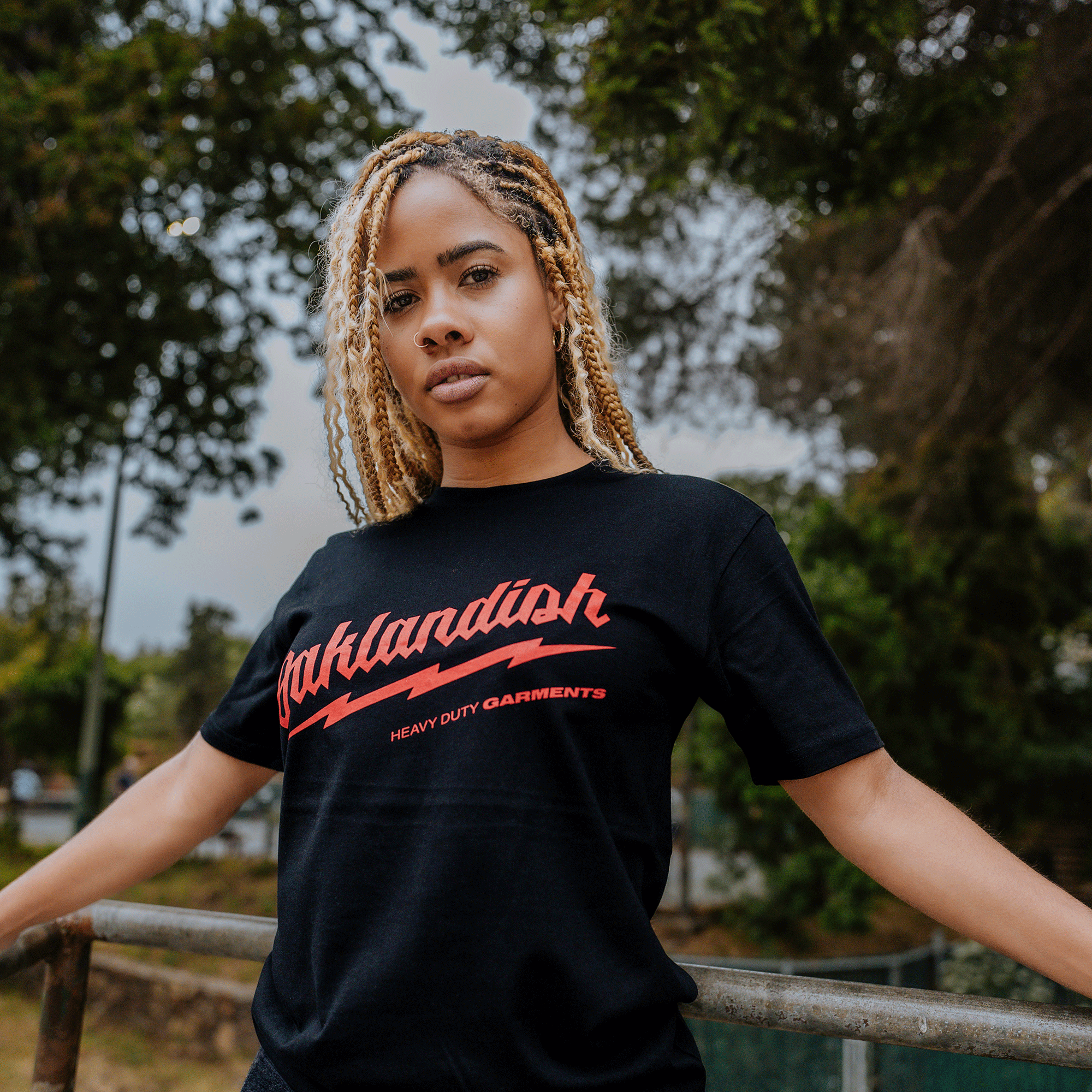 A person with braided blonde hair is standing outdoors, wearing the Oaklandish Power Tools Tee, a black T-shirt made of 100% cotton. The shirt features the word "Oaklandish" in red script across the front, followed by "Heavy Duty Garments" in smaller red font. They are leaning against a metal railing, with trees and greenery in the background.