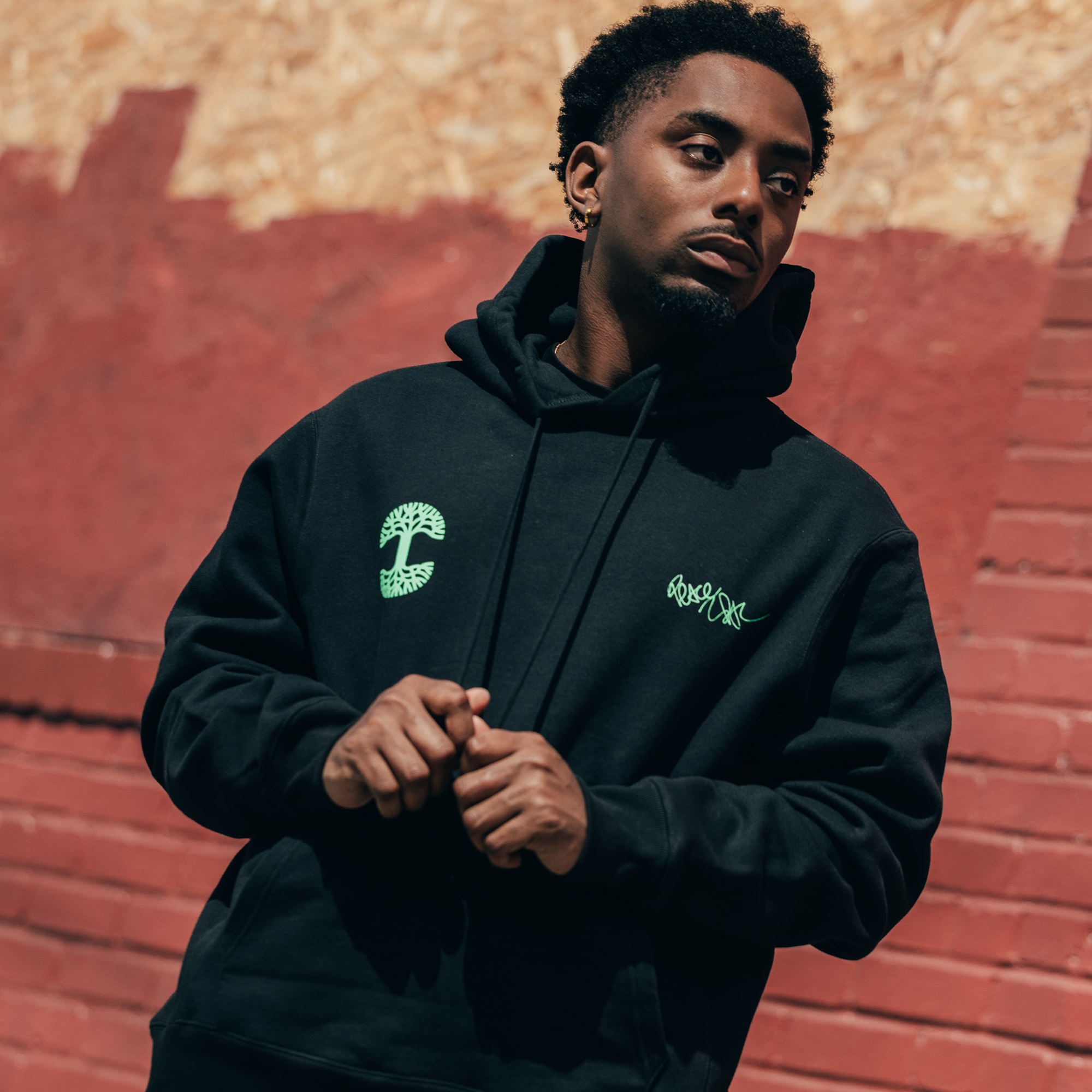 In front of a red brick wall, a person wearing the Oaklandish You Got To KYHU Hoodie, adorned with green designs, exudes the essence of Oakland culture. Their short curly hair and serious expression perfectly capture the spirit of an Oaklandish collaboration. The vibrant lighting accentuates the contrasting beige and red backdrop.