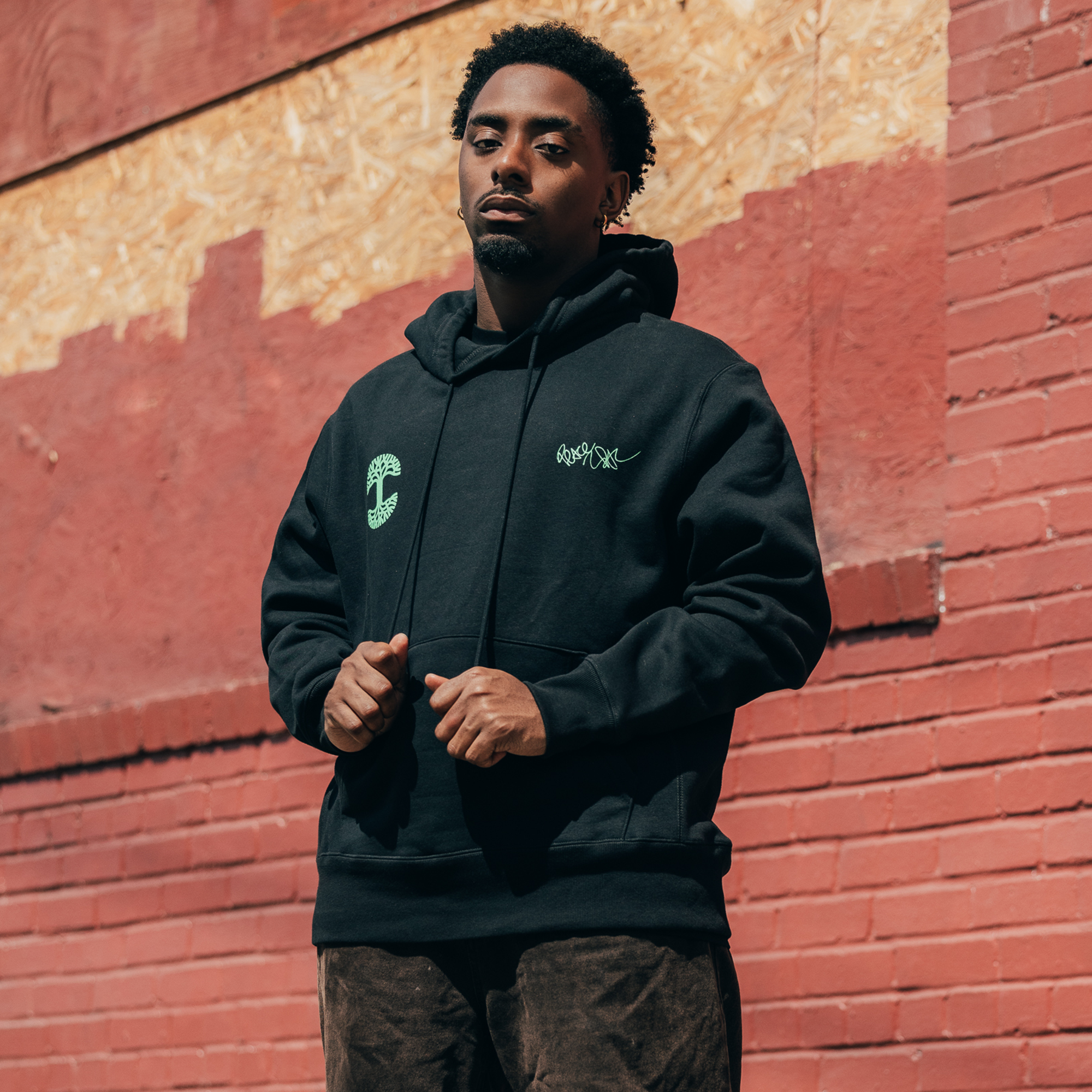 A person wearing the "You Got To KYHU Hoodie" from the Oaklandish x Tupac collaboration stands against a textured red brick wall with plywood above. They have short curly hair and are looking forward with a serious expression. The sunny background casts a shadow on the wall.