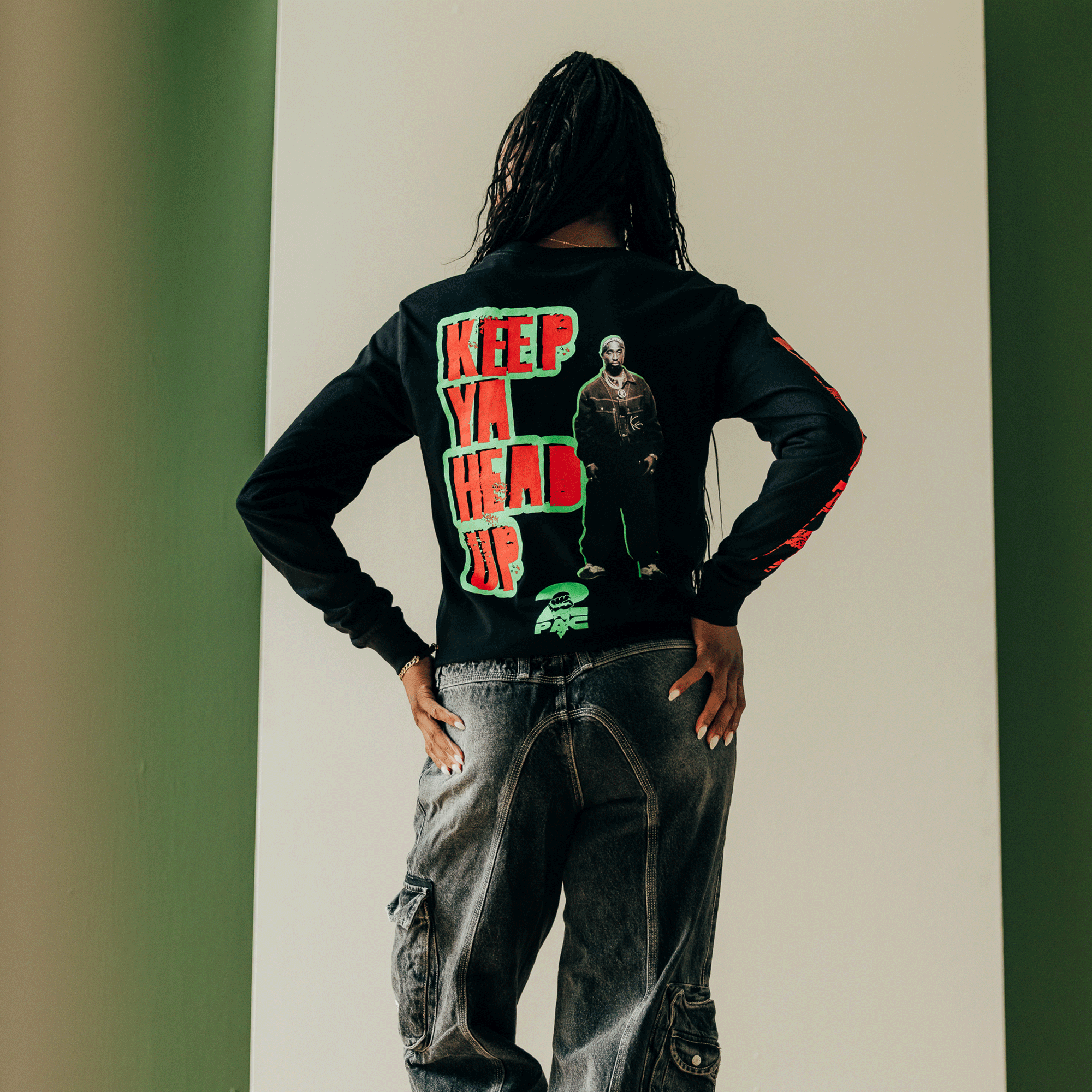 A person with long braided hair stands against a white and green wall, wearing the "You Got To KYHU LS Tee" from the Oaklandish collection. This black long-sleeve tee, featuring bold red text and an illustrated image celebrating Tupac's cultural influence in Oakland, displays the message "KEEP YA HEAD UP." They pair it with dark, faded jeans.