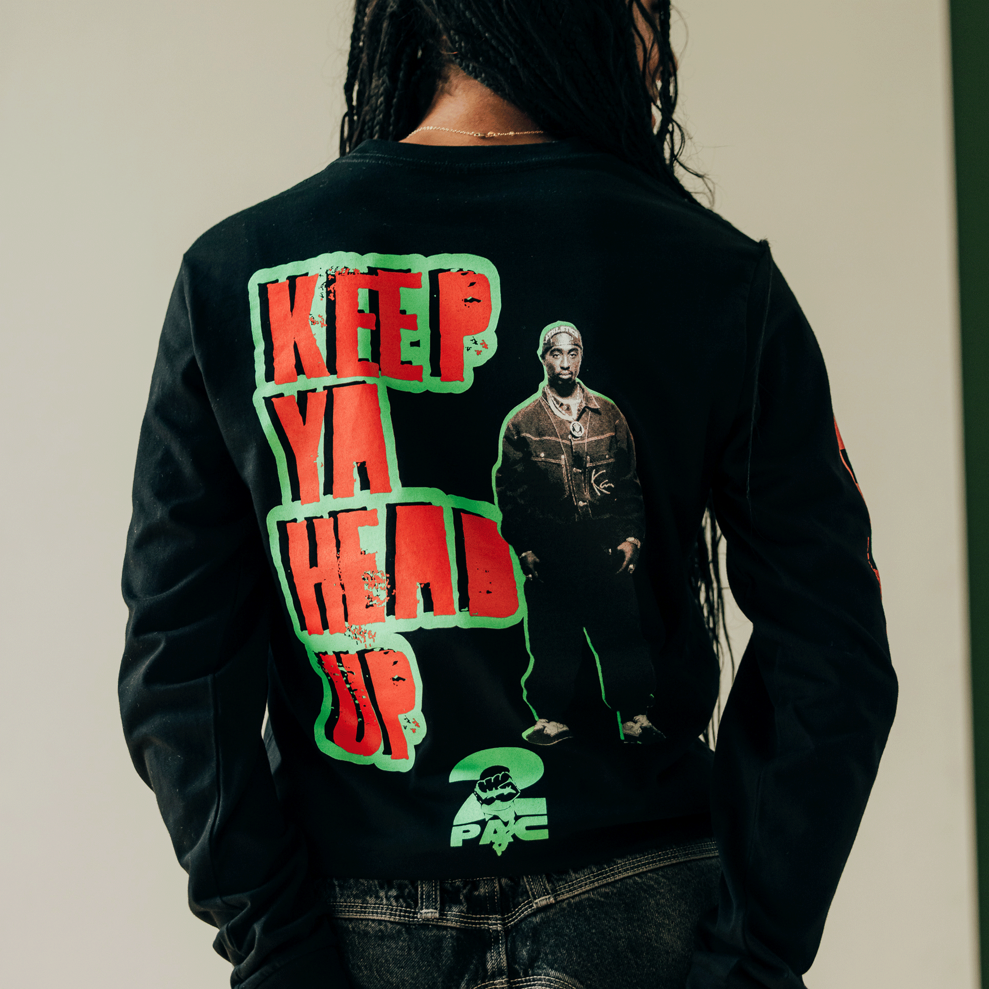 Someone with long braids stands facing away, sporting the You Got To KYHU LS Tee by Oaklandish. The black long-sleeve shirt showcases a design from the Oaklandish x Tupac collaboration, featuring bold red and green "KEEP YA HEAD UP" text beside a striking image of a man in a leather jacket on the back.