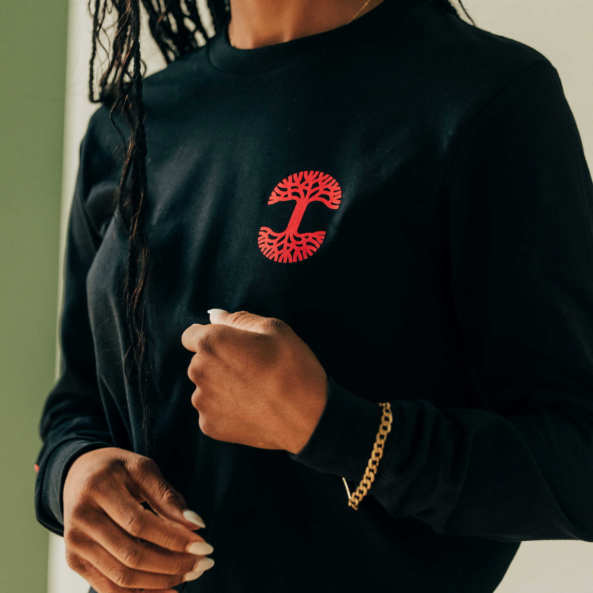 A person with long braided hair wears the Oaklandish "You Got To KYHU LS Tee," which features a red tree and Tupac design on the chest. They hold the fabric with one hand, showcasing white painted nails, while a gold chain bracelet adorns their wrist. The softly lit background creates a serene ambiance.