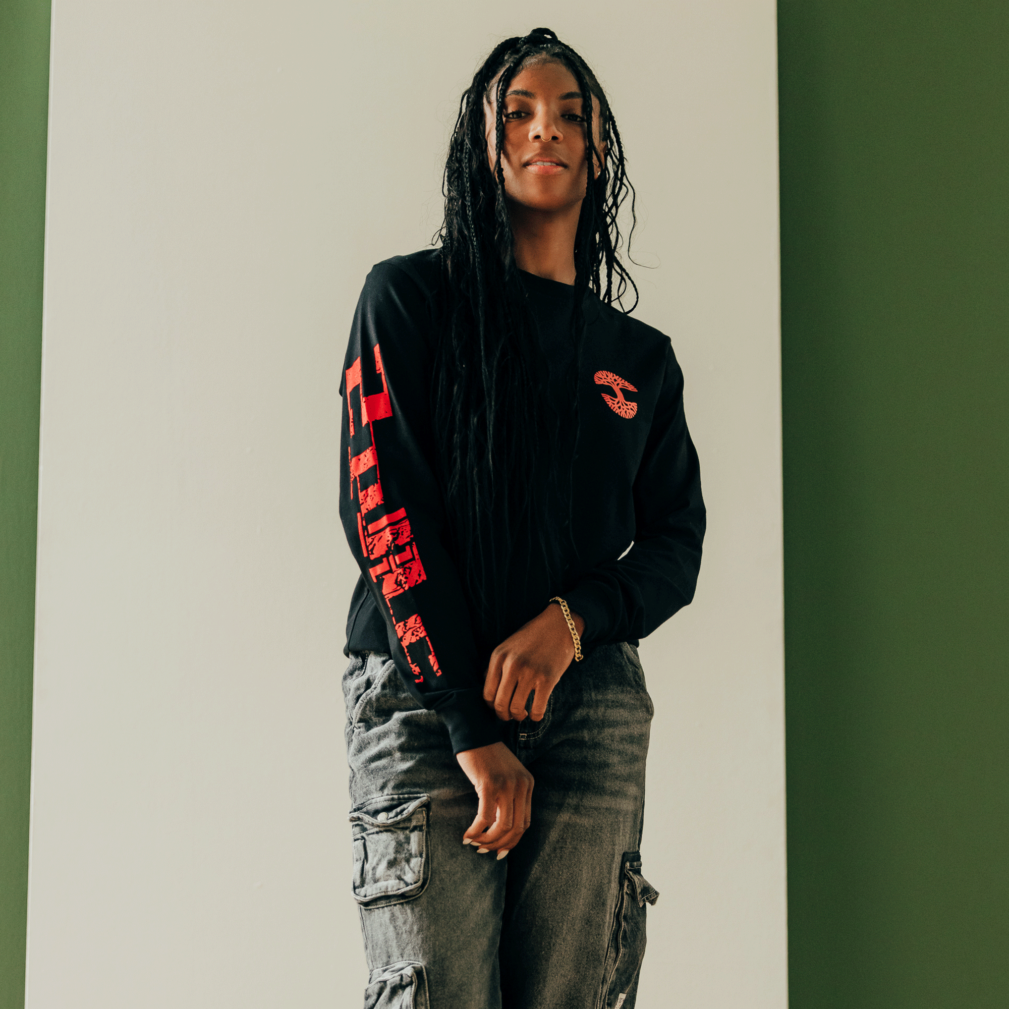 A person with long braided hair stands confidently against a green and white wall, wearing the Oaklandish "You Got To KYHU" long sleeve tee, featuring red lettering on the sleeve and a red graphic on the chest. They sport gray cargo pants and accessorize with bracelets, embodying Oakland's cultural impact.